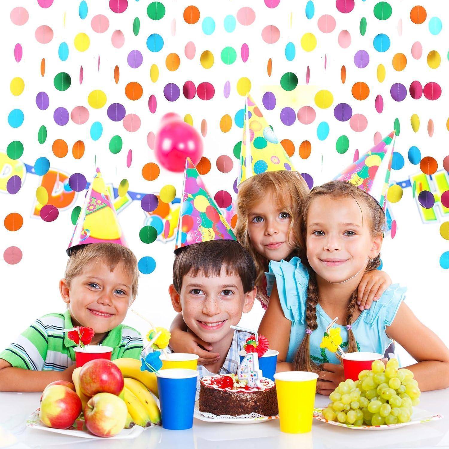 Vier Kinder mit Partyhüten lächeln an einem Geburtstagstisch mit Kuchen, Obst, Getränken und Trauben vor einem bunten Konfetti-Hintergrund.