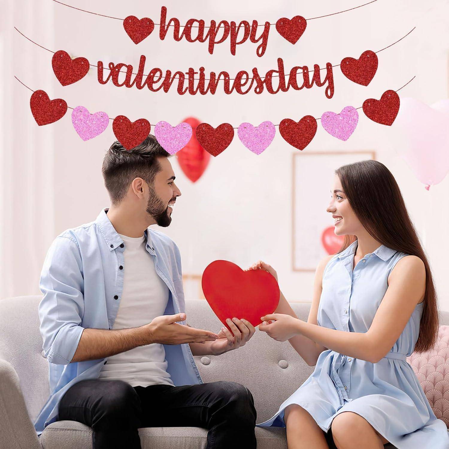 Ein lächelndes Paar sitzt auf einem Sofa und tauscht eine rote herzförmige Karte aus, mit einem „Alles Gute zum Valentinstag"-Banner und Herzgirlanden darüber.