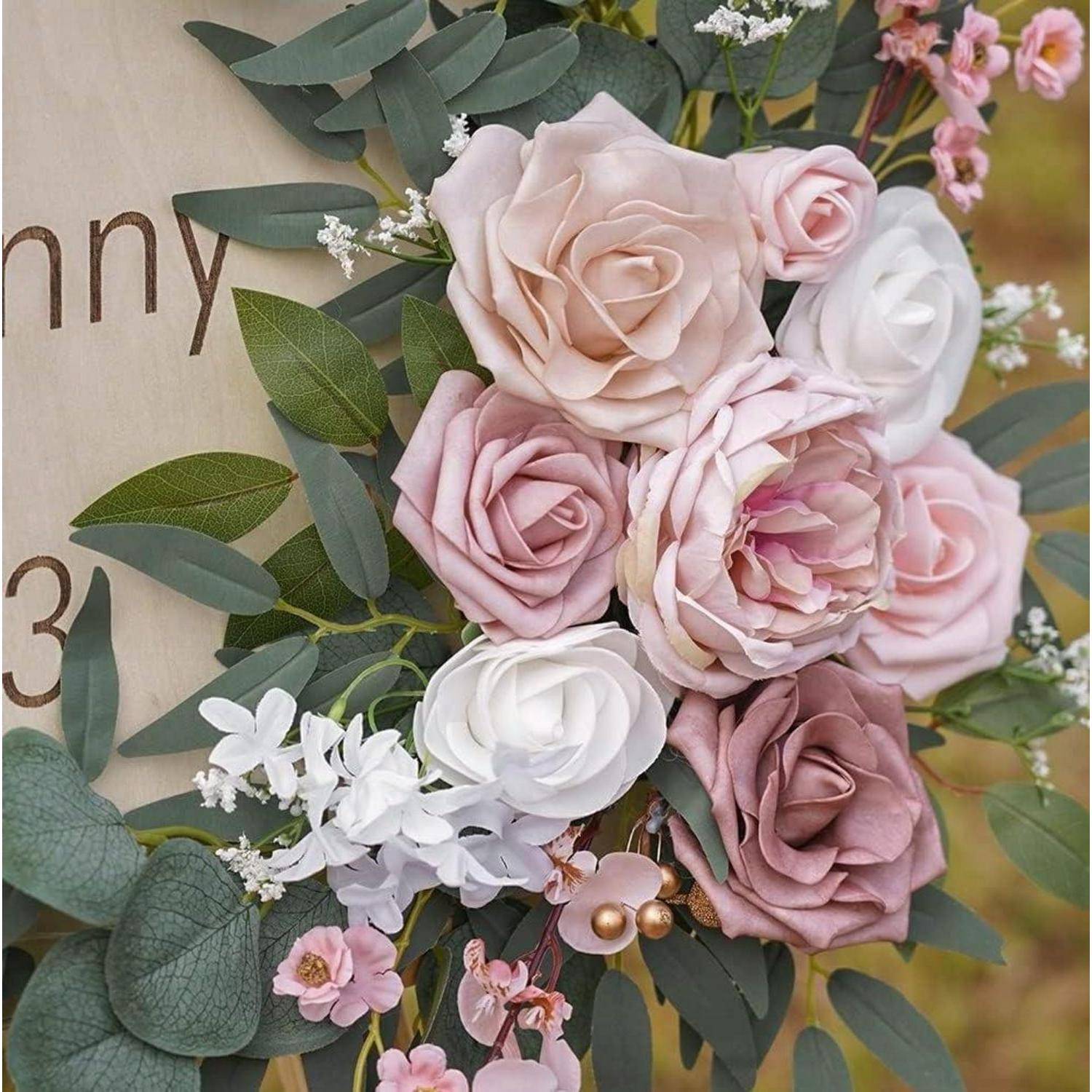 Nahaufnahme einer pastellfarbenen Blumenarrangement mit rosa, puderrosa und weißen Rosen, grünen Eukalyptusblättern und kleinen weißen Füllblüten auf einem Schild oder Hintergrund.