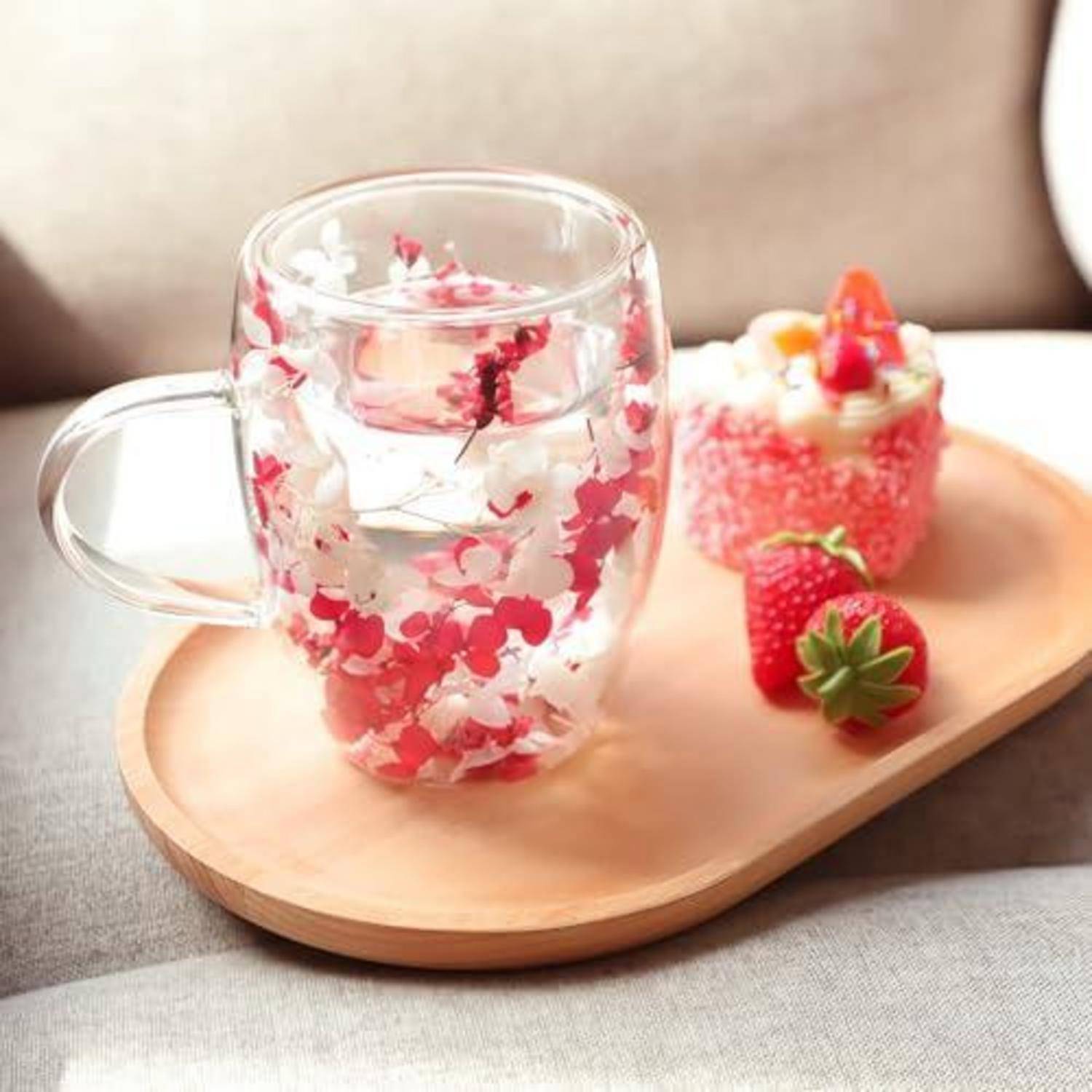Klare Glastasse mit rosa-weißem Blumenmuster auf einem Holztablett, neben einem Erdbeer-Dessert und frischen Erdbeeren auf einem Sofa.