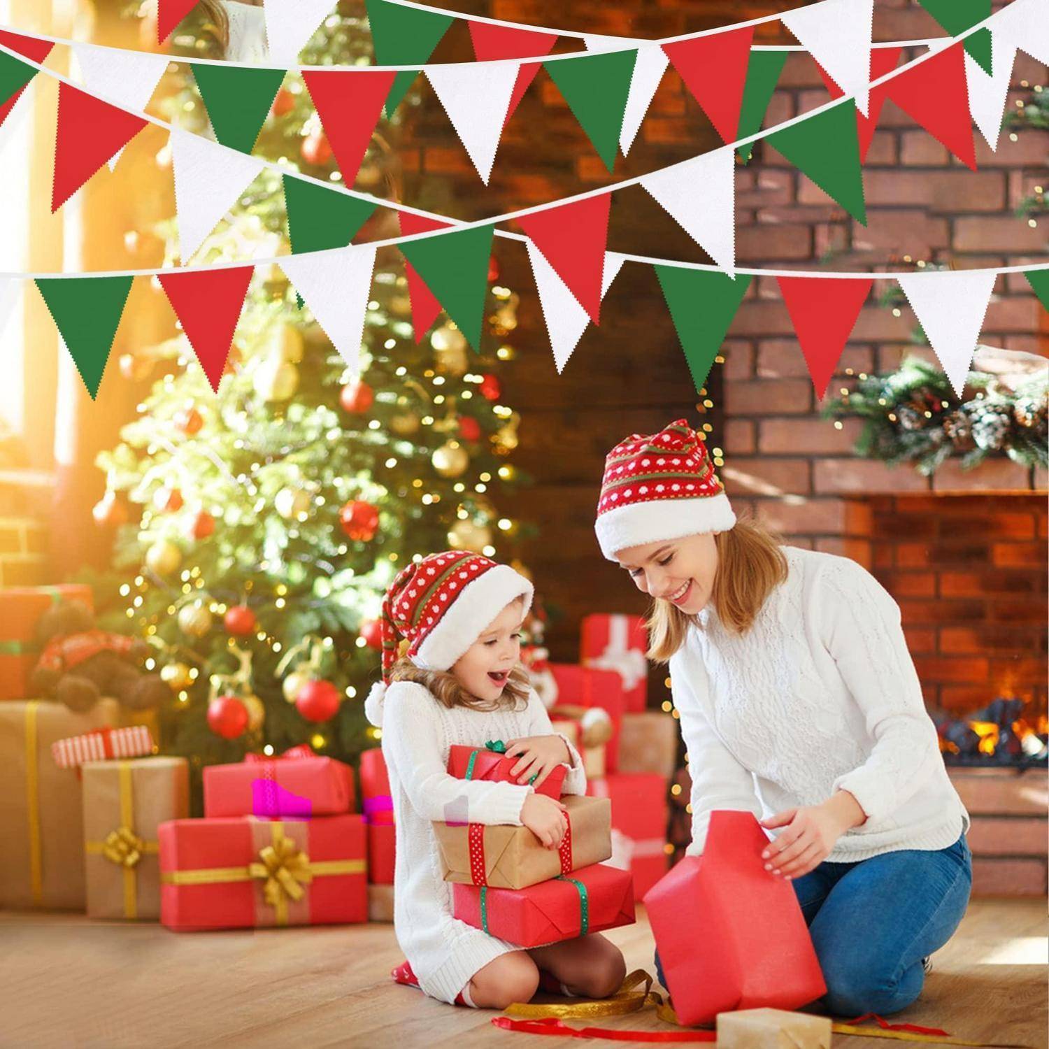 Frau und junges Mädchen in Weihnachtsmützen öffnen Weihnachtsgeschenke neben einem geschmückten Baum und Kamin, mit roter, grüner und weißer Girlande oben.