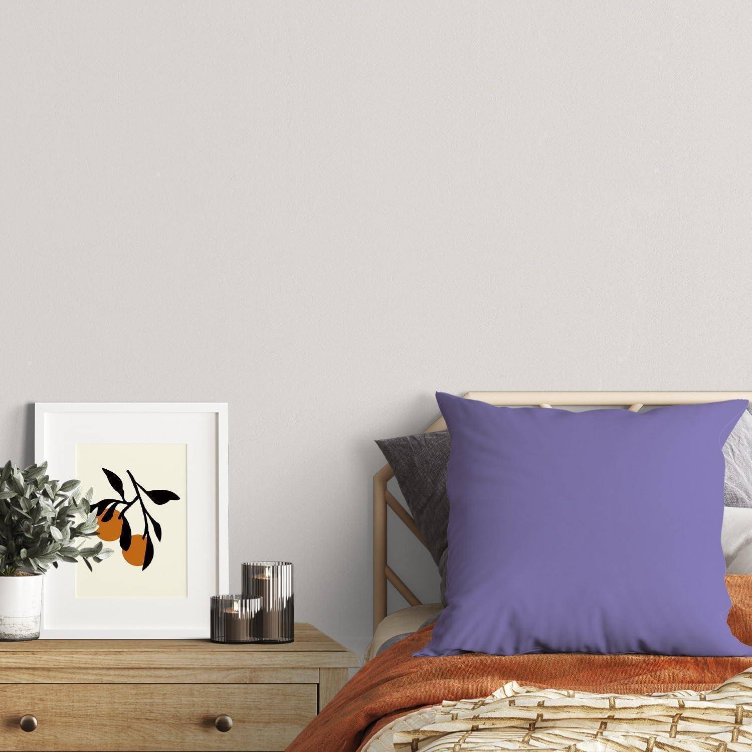 Schlafzimmerszene mit einem Bett, einem lila Kissen, einer orangefarbenen Decke und einem Nachttisch, auf dem sich gerahmte Kunstwerke, eine Pflanze und Kerzen befinden.