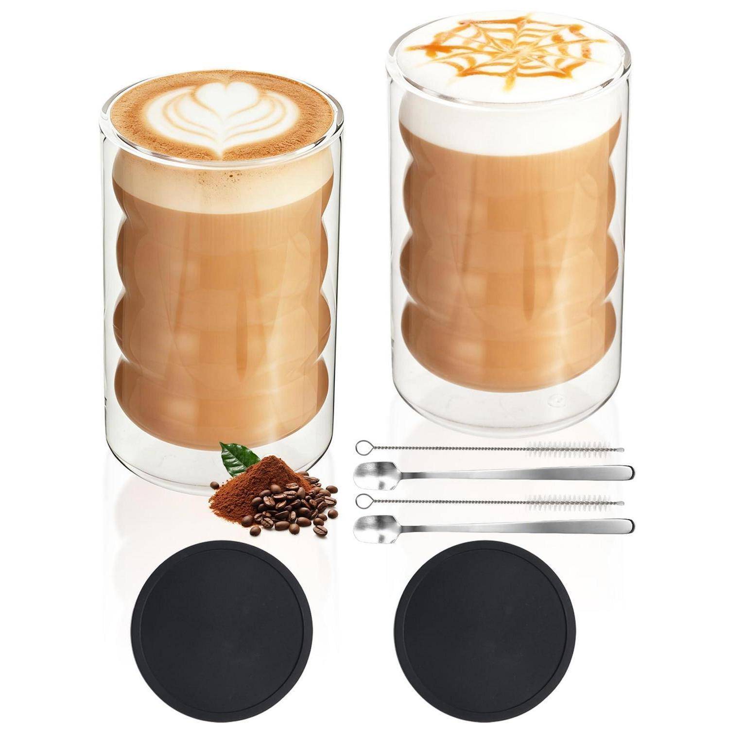 Zwei klare doppelwandige Kaffee-Gläser mit Latte Art, gezeigt mit zwei schwarzen Untersetzer, Kaffeebohnen und zwei Metallöffel.