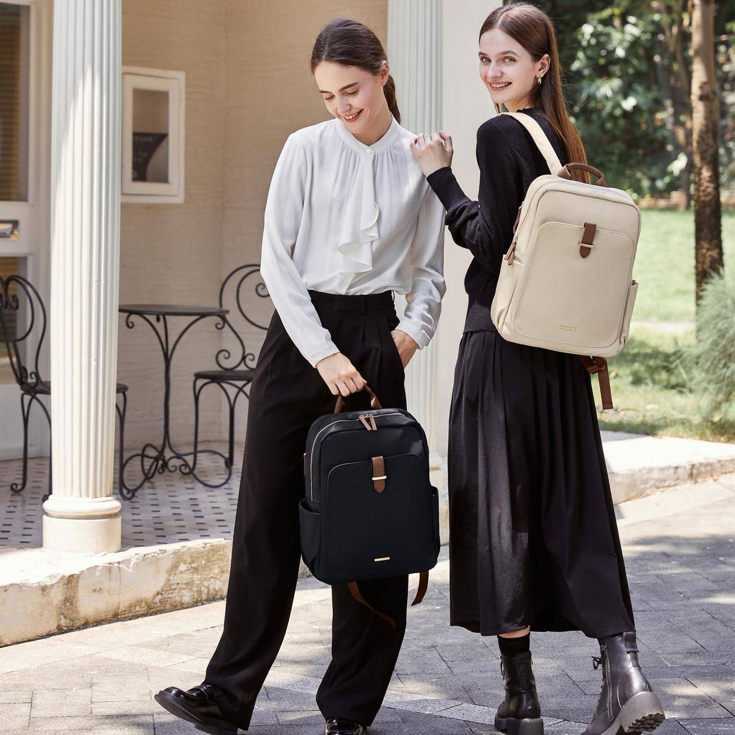 Zwei Frauen in Schwarz-Weiß-Outfits stehen draußen mit Rucksäcken, eine hält eine schwarze Tasche und die andere trägt einen beigen Rucksack, während sie in die Kamera lächeln.