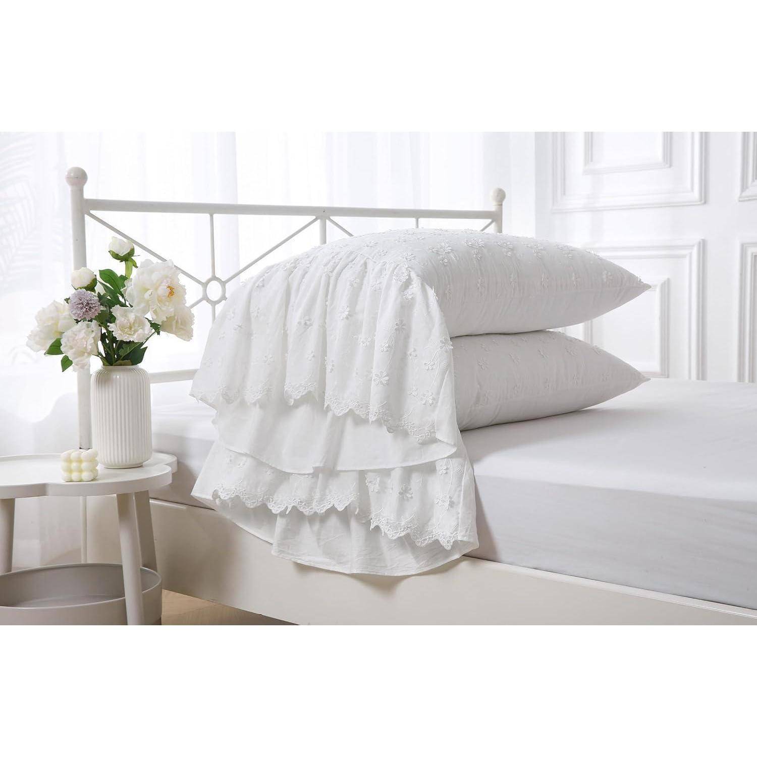 Ein ordentlich gemachtes weißes Bett mit aufgestapelten Kissen, einer weiß gekante Decke mit Spitzensaum und eine Vase mit Blumen auf einem Nachttisch in einem hellen Schlafzimmer.