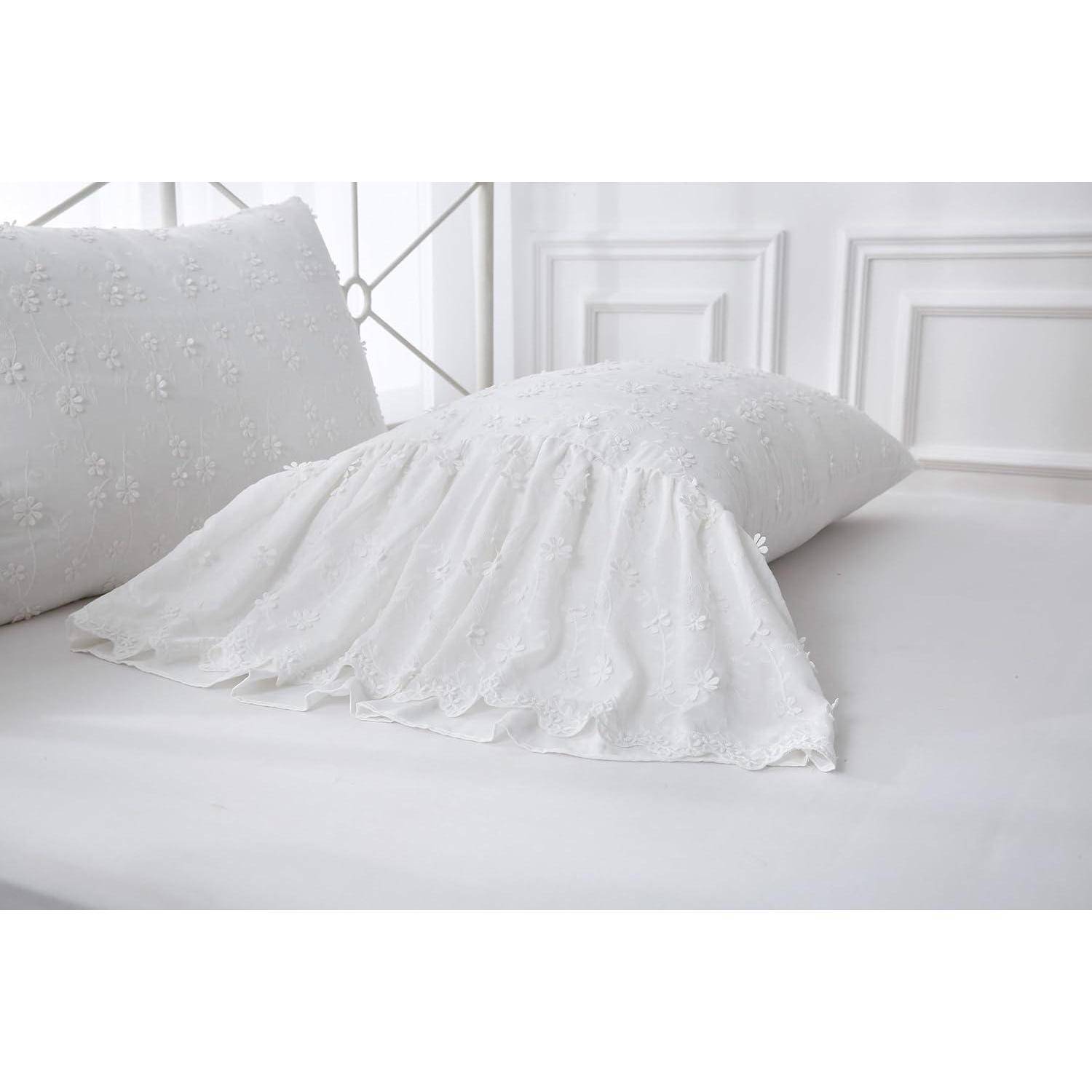 Weißes Kissen mit Spitzenbesatz auf einem Bett mit passendem besticktem Kissen, gezeigt in einem hellweißen Schlafzimmer.