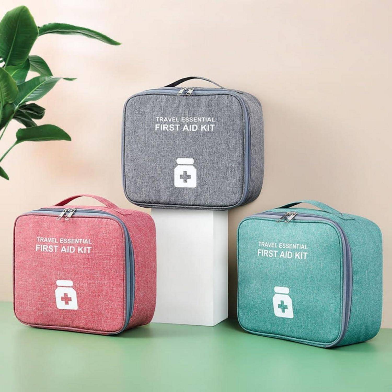 Drei Reise-Erste-Hilfe-Kits mit Reißverschluss in Grau, Rosa und Türkis, jeweils mit der Beschriftung „TRAVEL ESSENTIAL FIRST AID KIT" und einem medizinischen Kreuz-Symbol.
