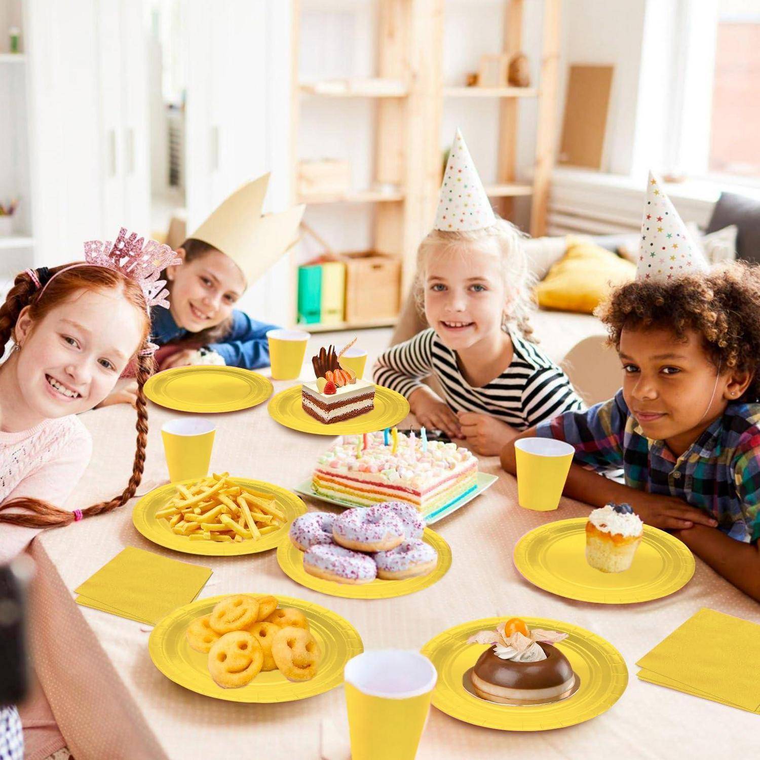 Vier Kinder mit Partyhüten lächeln um einen Tisch herum, auf dem Geburtstagskuchen, Cupcakes, Donuts, Pommes frites und gelbe Teller und Tassen stehen.