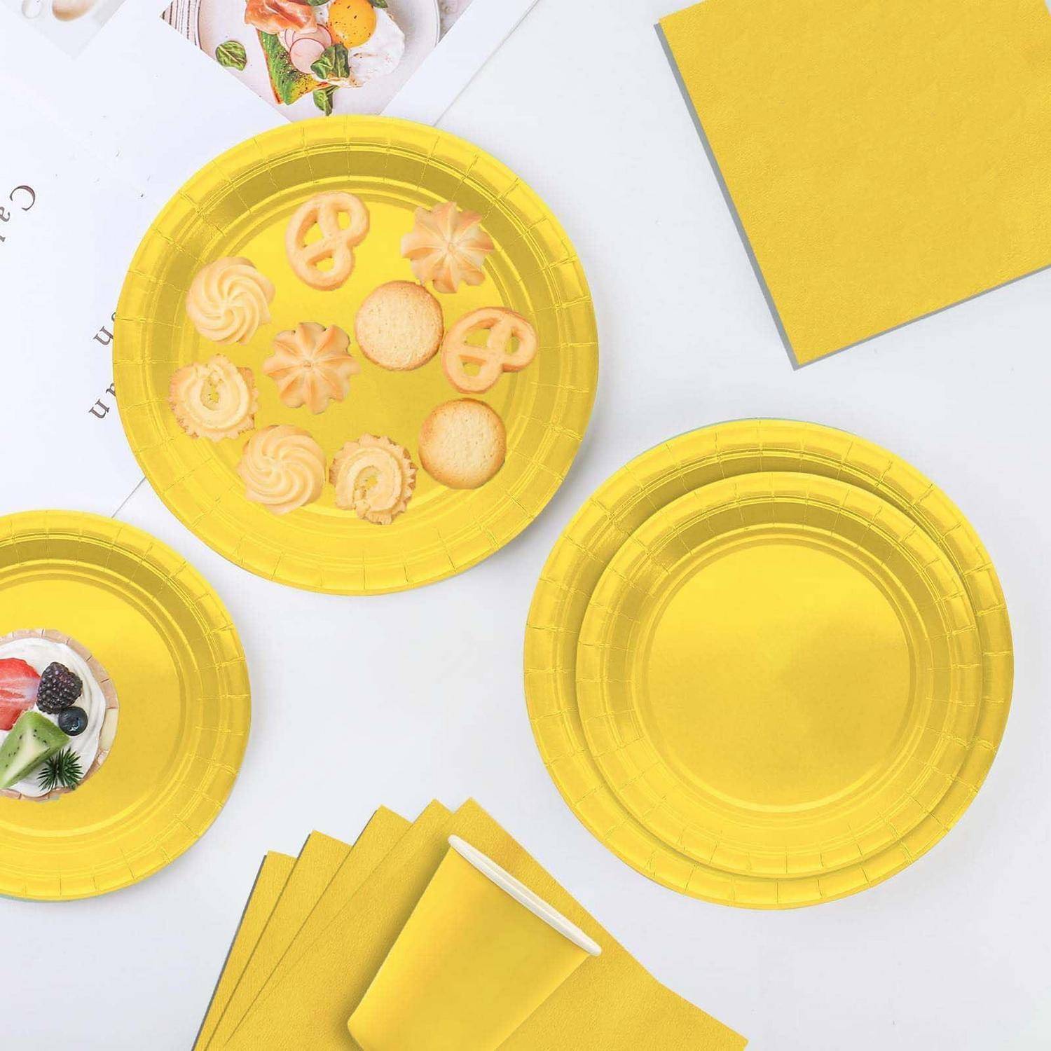 Gelbe Einwegplatten und Servietten auf einer weißen Oberfläche, wobei ein Teller assortierte Kekse hält und ein anderer ein Dessert mit Beeren.