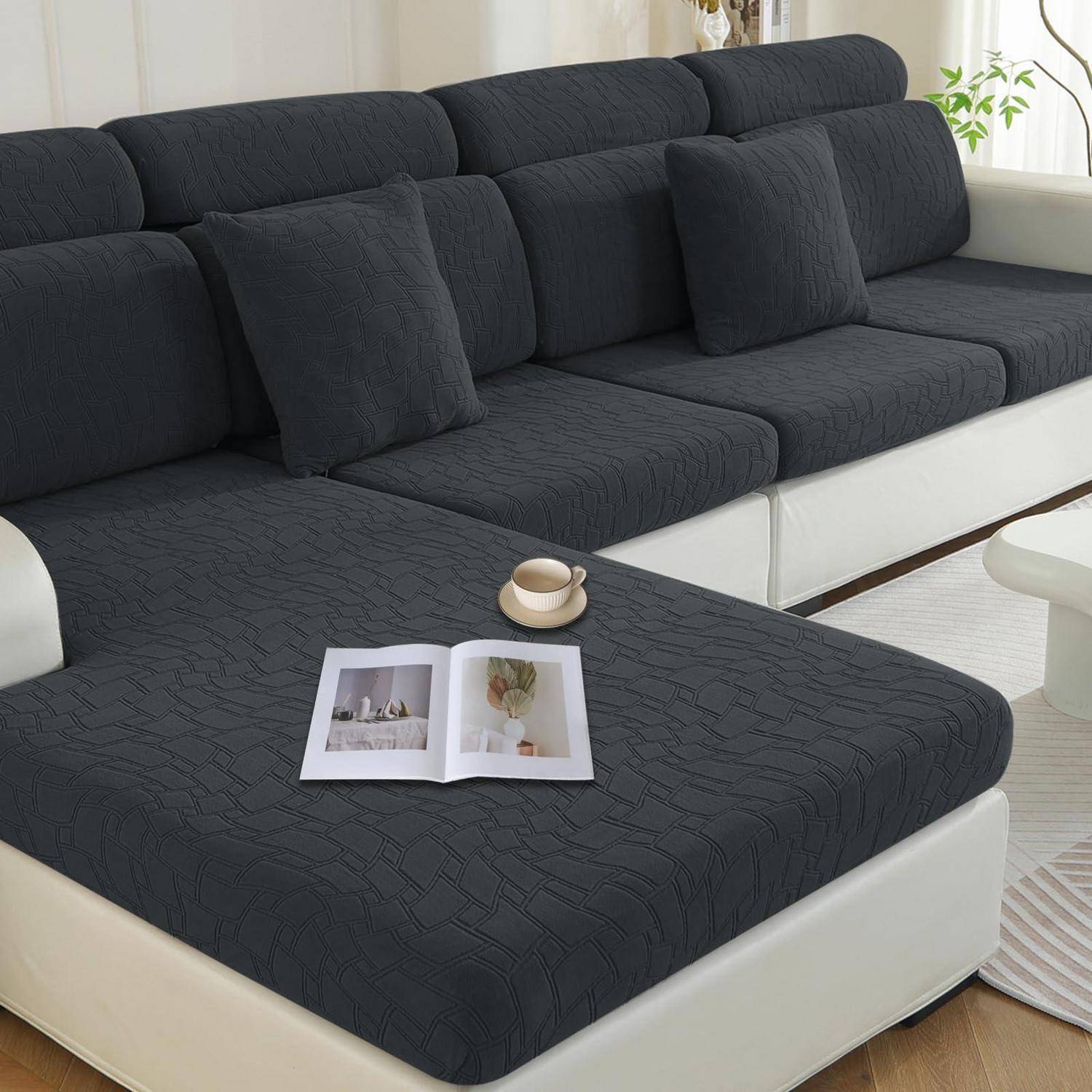 Ein dunkelgraues Ecksofa mit weißem Sockel und Kissen, mit einer offenen Zeitschrift und einer Tasse auf dem Hocker; helle Wohnzimmerumgebung.