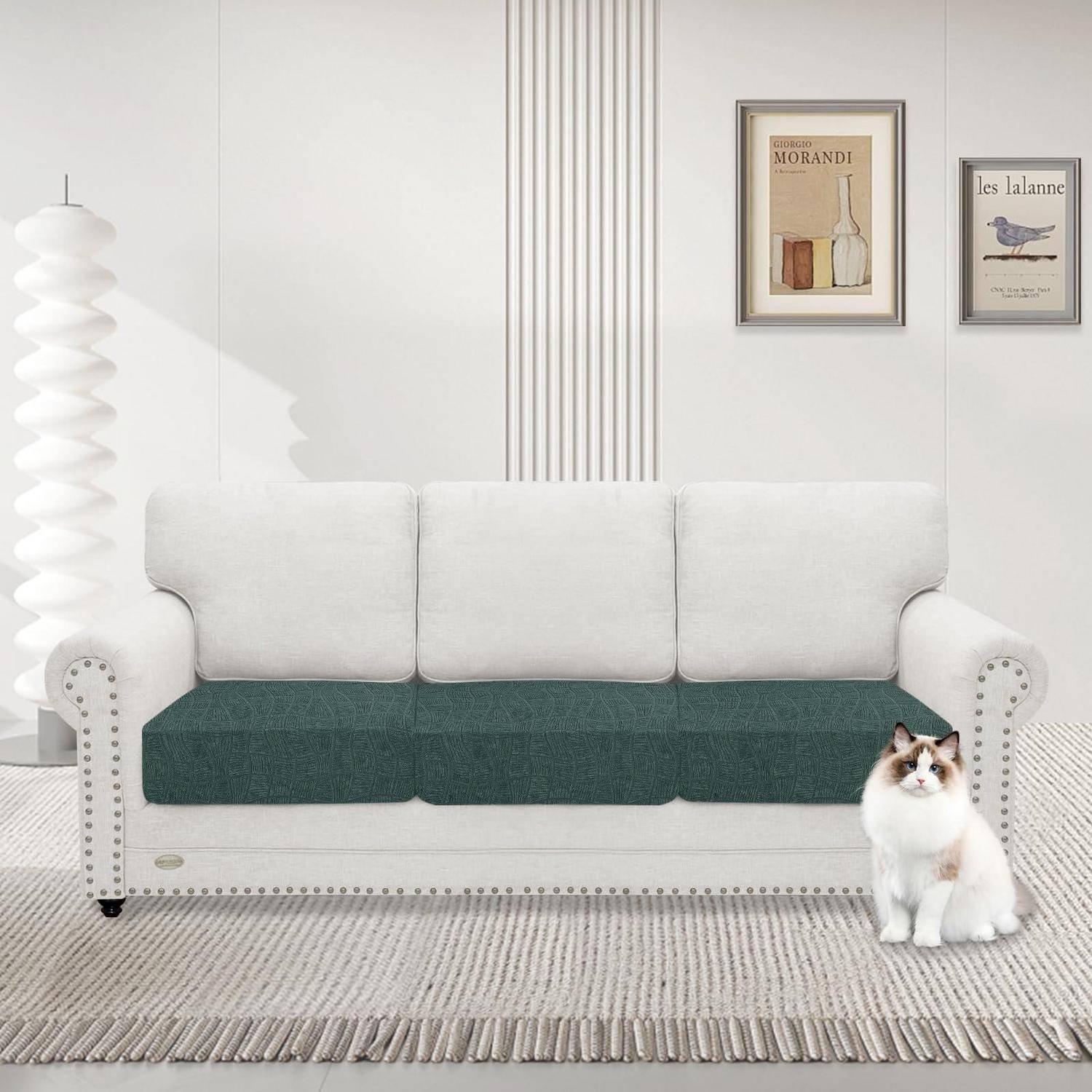 Weiße Chesterfield-Sofa mit grünen Sitzkissen in einem hellen minimalistischen Raum, mit gerahmten Wandbildern, einer skulpturalen Stehleuchte und einer Katze, die daneben sitzt.