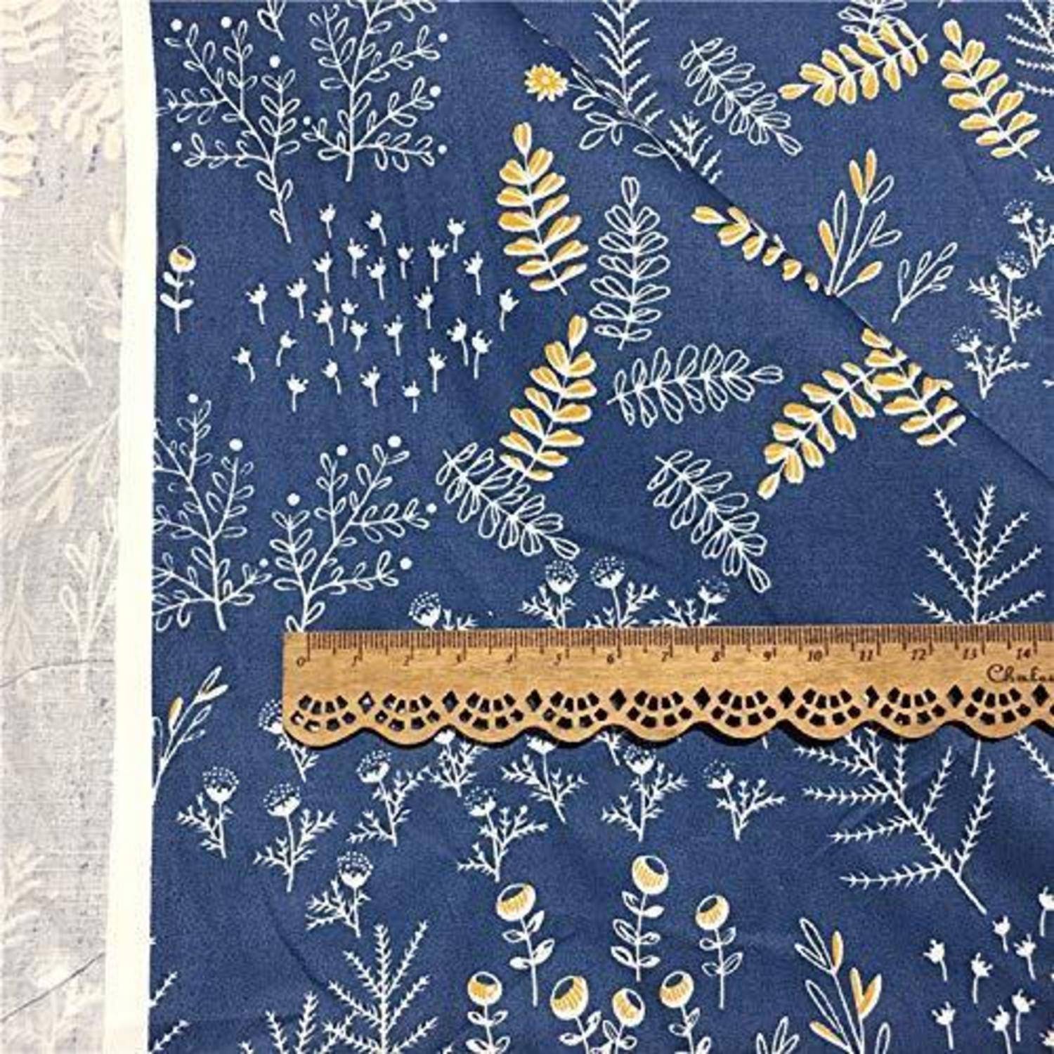 Nahaufnahme von blauem Stoff mit Muster, weißen und gelben botanischen Drucken, ein dekoratives Spitzenlineal in der Mitte zur Größenangabe.