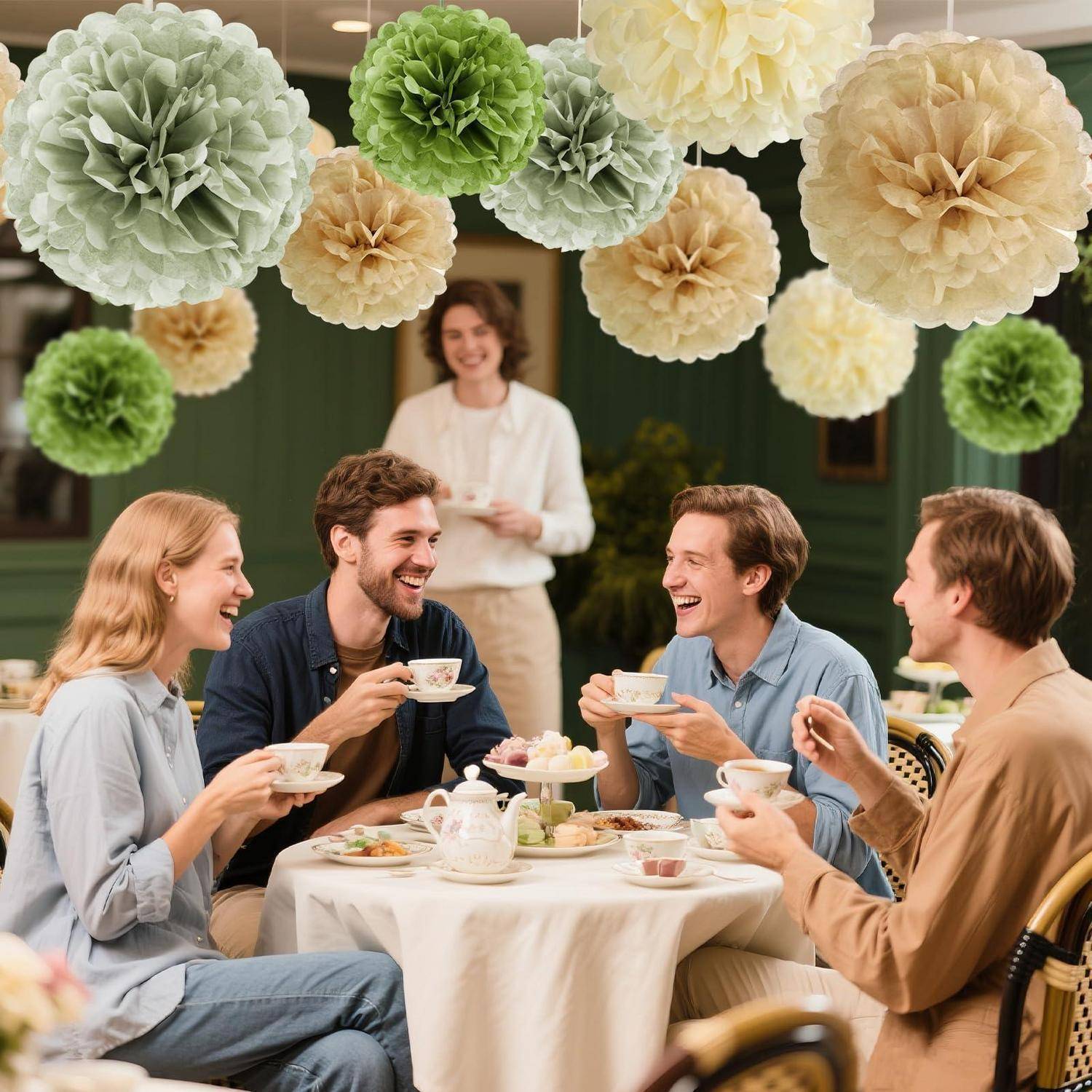 Vier Freunde unterhalten sich lachend bei Tee an einem kleinen runden Tisch in einem dekorierten Café, mit Papier-Pompoms als Blumendekoration über ihnen und einem Server im Hintergrund.