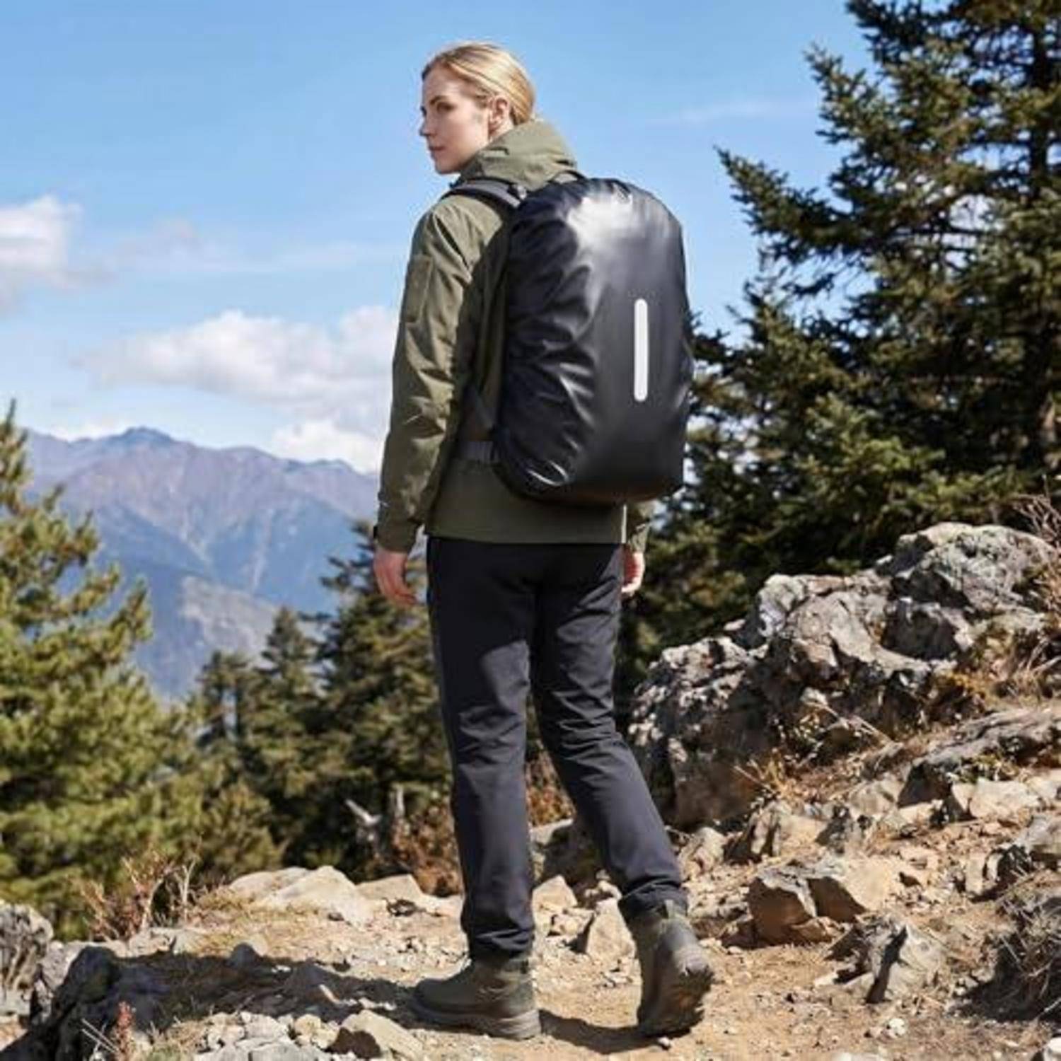 Eine Person wandert auf einem felsigen Bergpfad und trägt eine olivfarbene Jacke und einen schwarzen Rucksack mit einem reflektierenden Streifen.