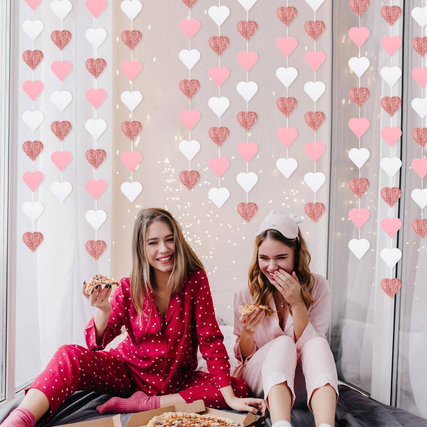 Zwei Frauen im Pyjama sitzen auf dem Boden, essen Pizza und lachen unter rosa, weißen und glitzernden Herzdekorationen.