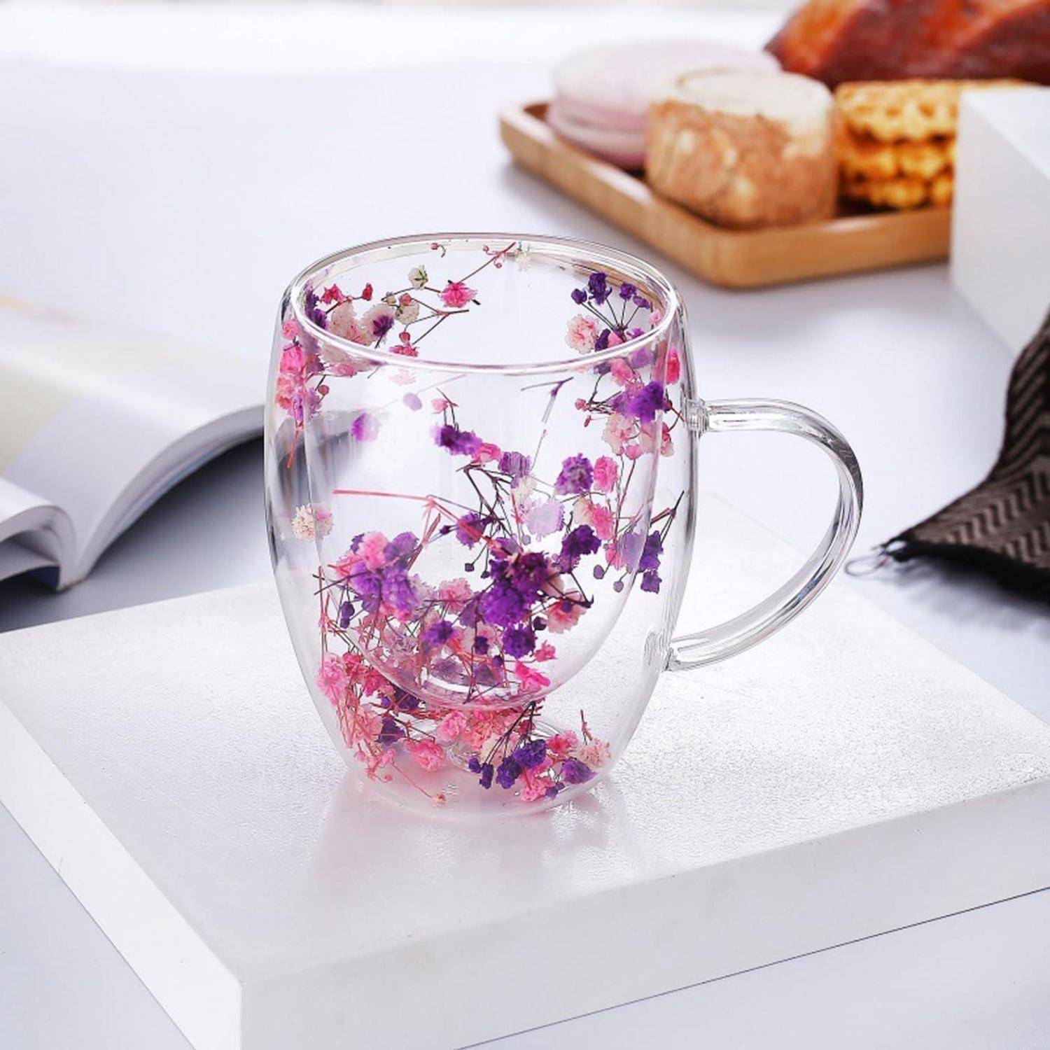 Transparente doppelwandige Glastasse gefüllt mit rosa und violetten Blumen, angezeigt auf einer weißen Oberfläche mit Büchern und Snacks im Hintergrund.
