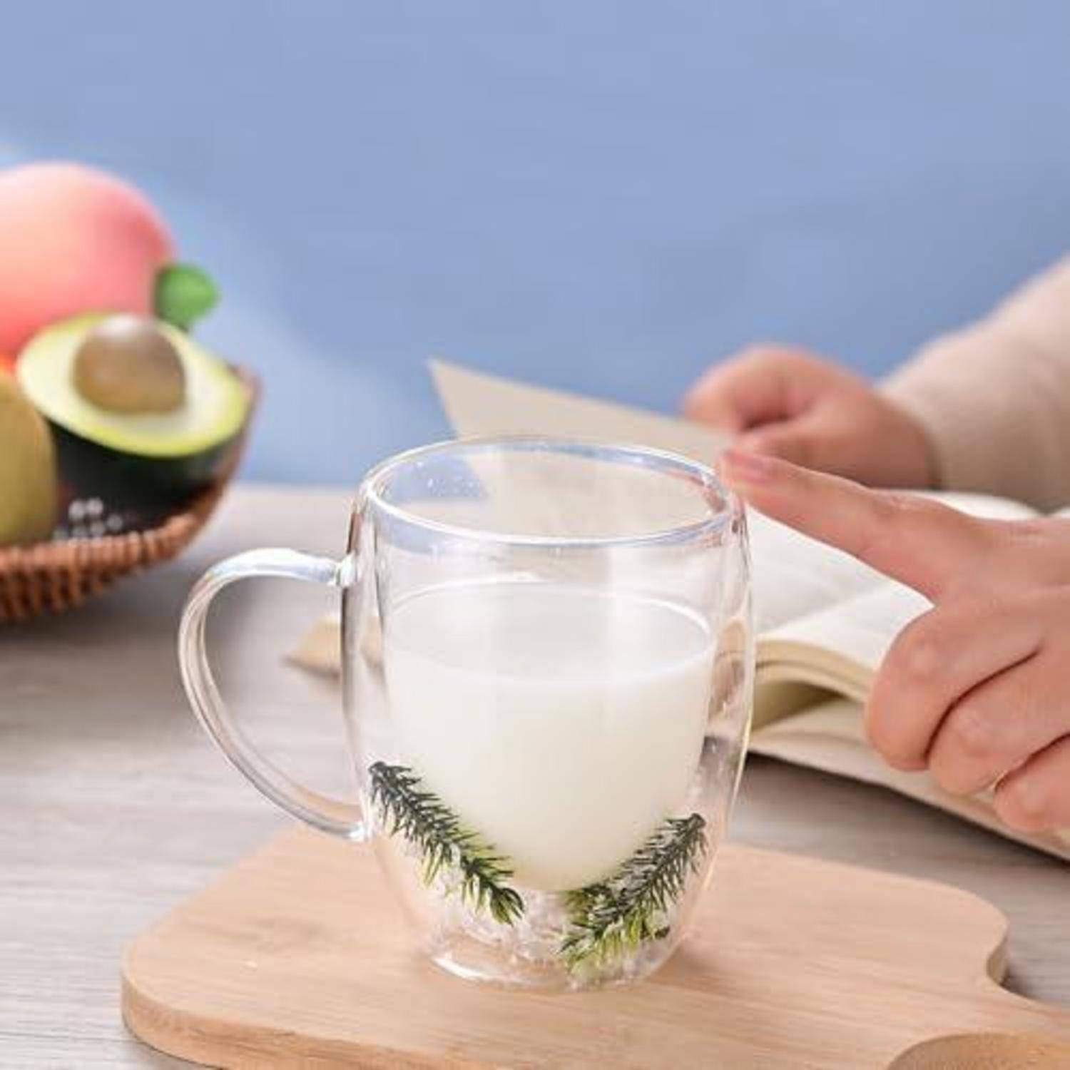 Eine Hand zeigt auf eine Glastasse mit Milch auf einem Holzbrett, mit einer Avocado und einem aufgeschlagenen Buch im Hintergrund.