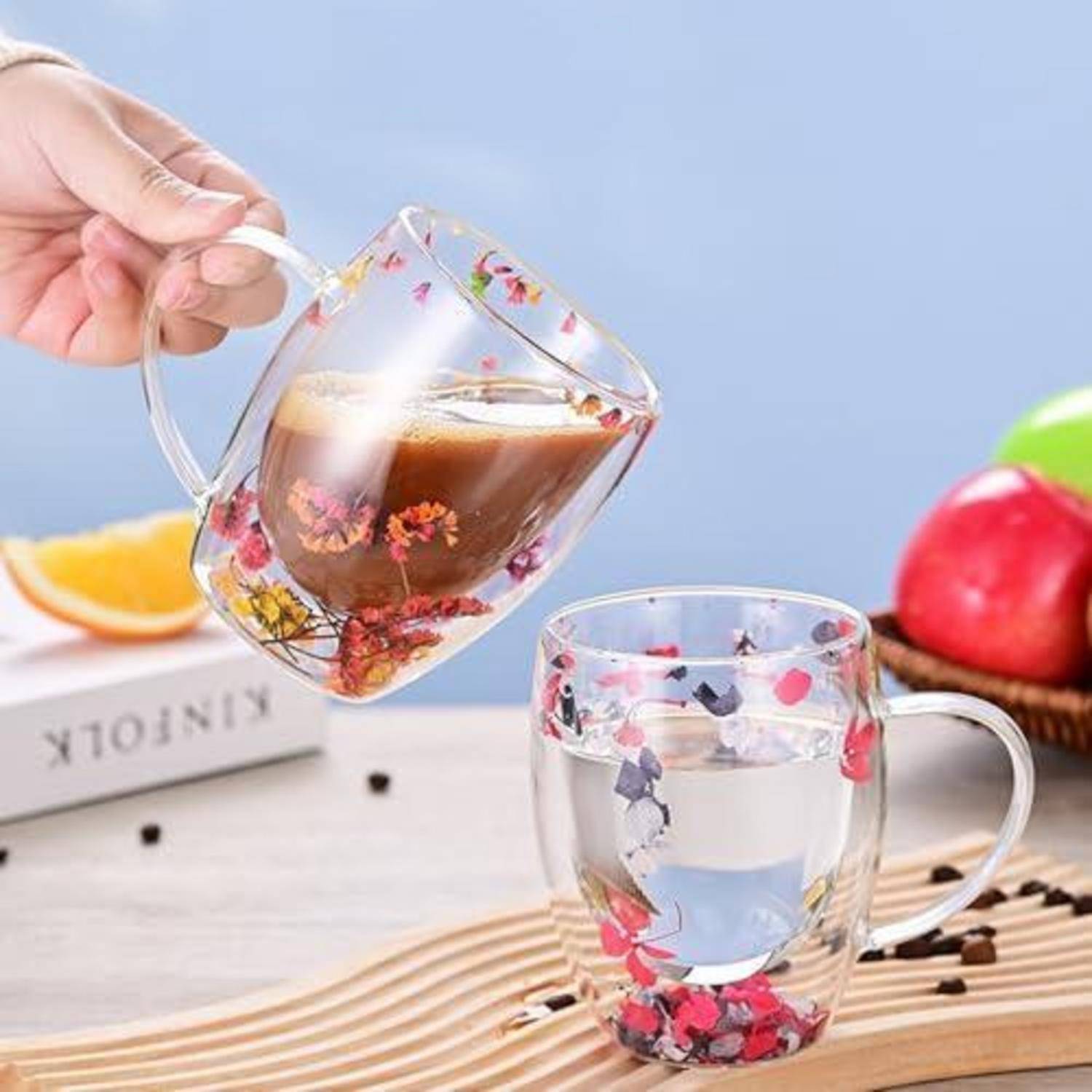 Eine Hand gießt Kaffee in eine florale doppelwandige Glastasse neben einer klaren passenden Tasse auf einem Tisch mit Obst und Kaffeebohnen.
