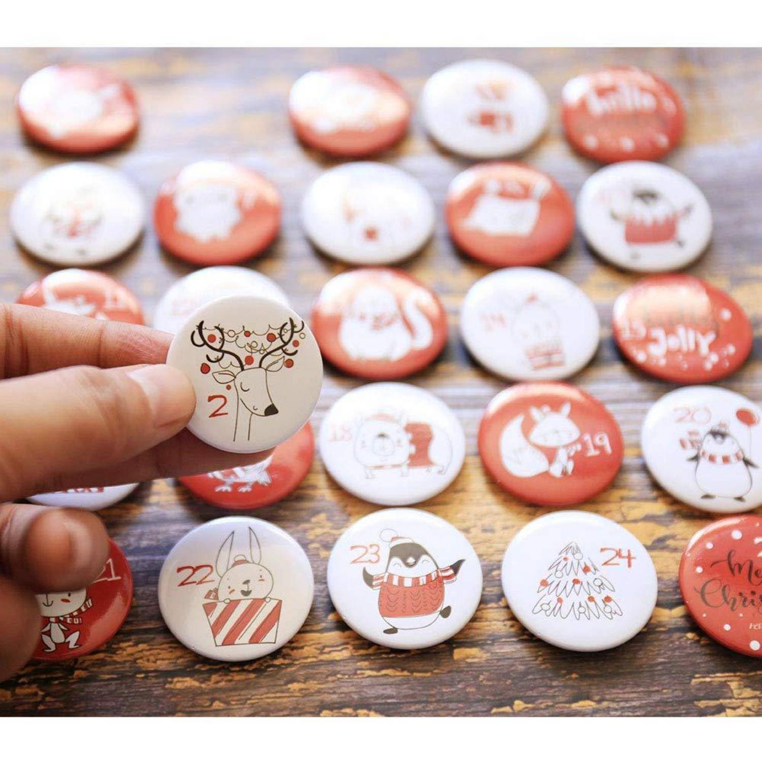 Eine Hand hält einen runden Adventskalender-Knopf mit der Markierung „2" mit einer Hirschillustration, umgeben von vielen nummerierten weihnachtsthematischen Knöpfen auf einer Holzoberfläche.