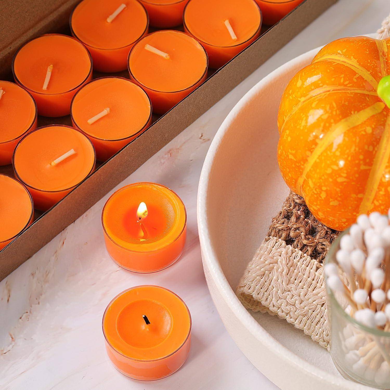 Orange Teelichter in einer Schachtel und auf einem Tisch neben einer weißen Schüssel, die einen kleinen dekorativen Kürbis und Herbstdeko aus Geflecht hält.