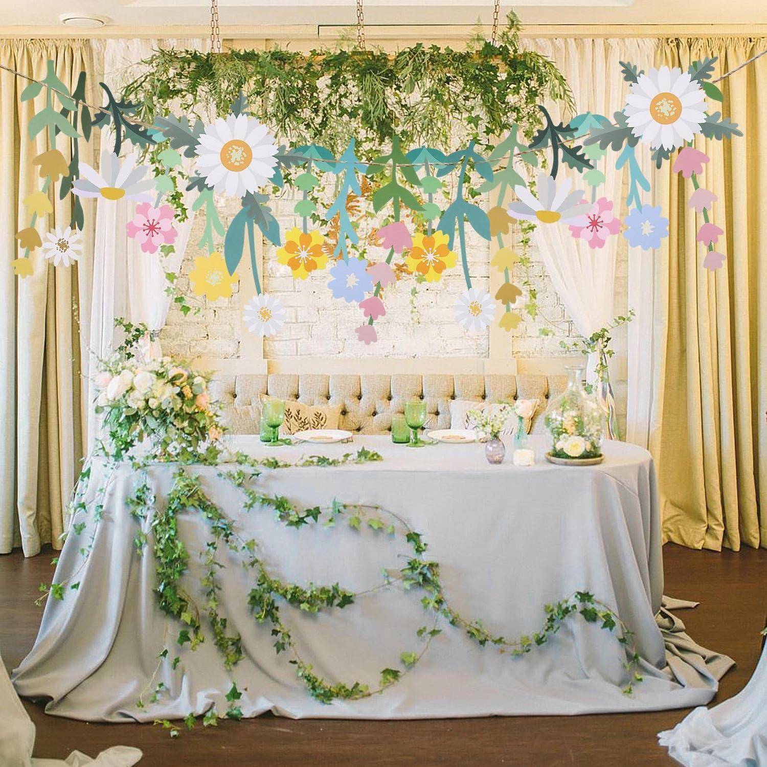 Dekorierter Partytisch mit weißem Tuch, Grüngirlande, Blumen und hängenden Papierblueten vor Vorhängen und einer gesteppten Kulisse.