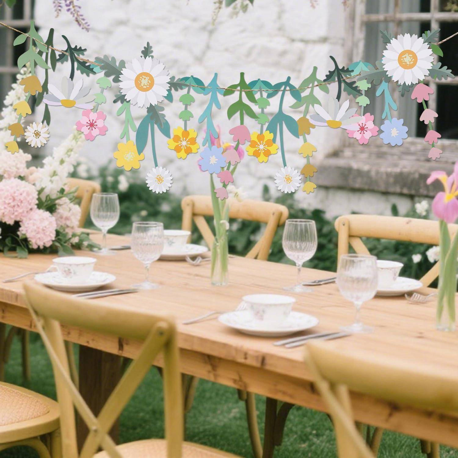 Dekorieter Außenesstisch mit Tellern, Gläsern und Blumen unter einer Girlande aus Papiertblüten in Pastellfarben, eingerichtet für eine Gartenfeier.