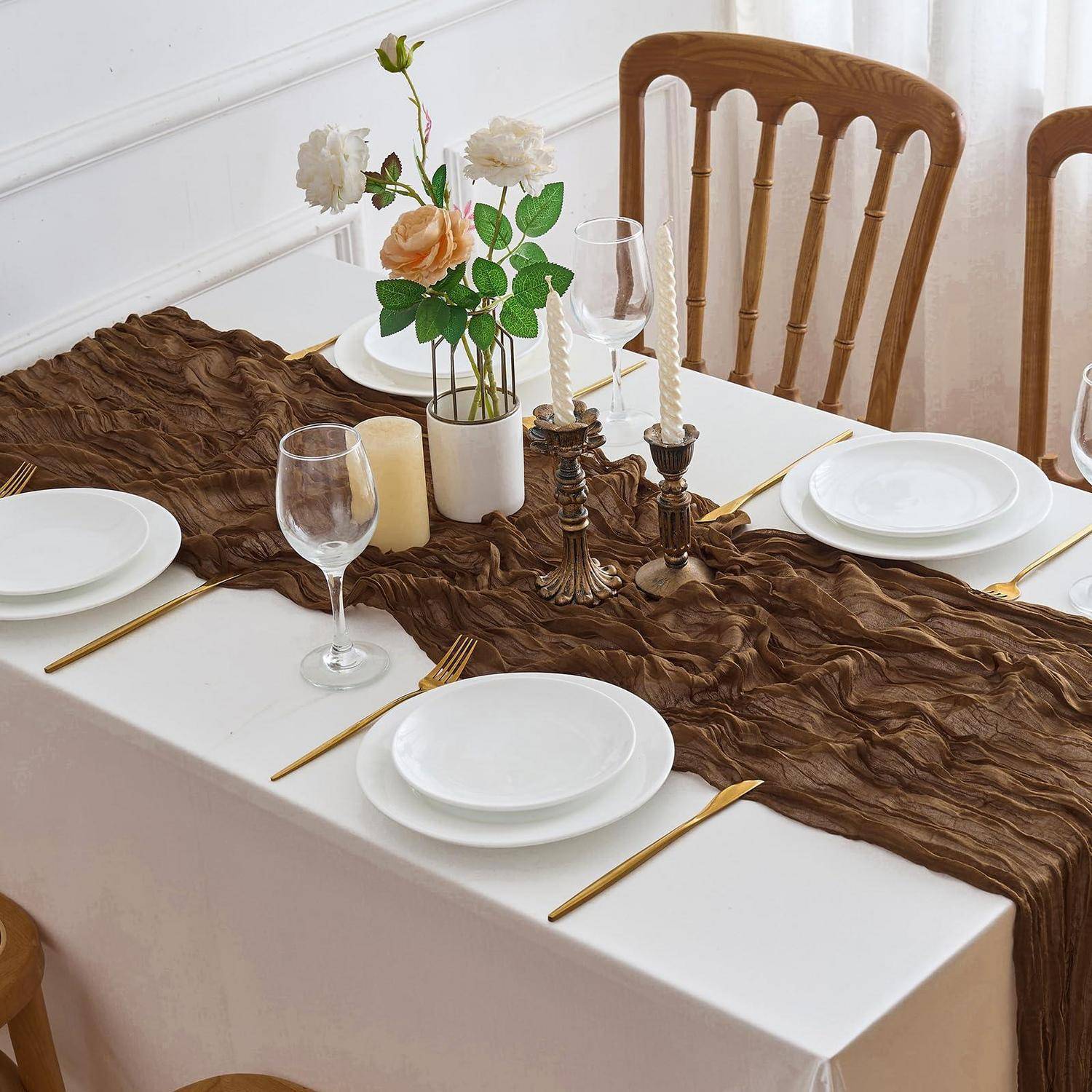 Formales Esszimmer für vier Personen mit weißen Tellern, goldenem Besteck, Weingläsern, Kerzen und einer Blumenmittelstückverzierung auf einem braunen strukturierten Läufer.