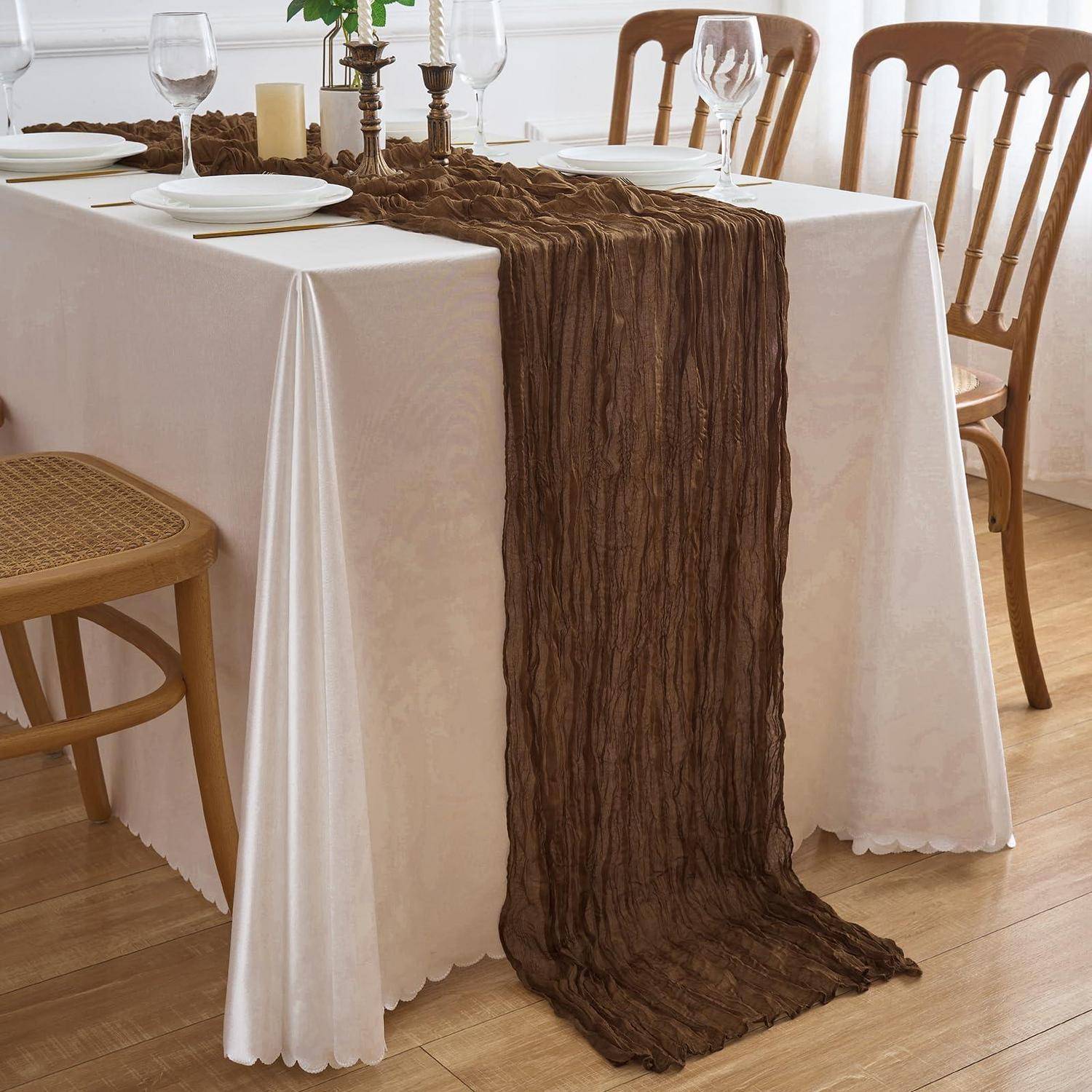 Esstisch mit weißer Tischdecke, Tellern, Gläsern und braunem strukturiertem Tischläufer, der über die Mitte und über die Kante drapiert ist, in einem hellen Raum mit Holzstühlen.