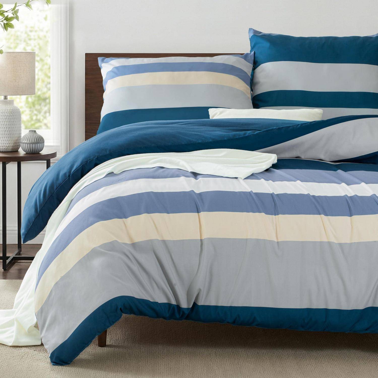 Gestreifter blau, creme und grauer Tagesdecke mit passenden Kissen auf einem ordentlich gemachten Bett in einem hellen Schlafzimmer.