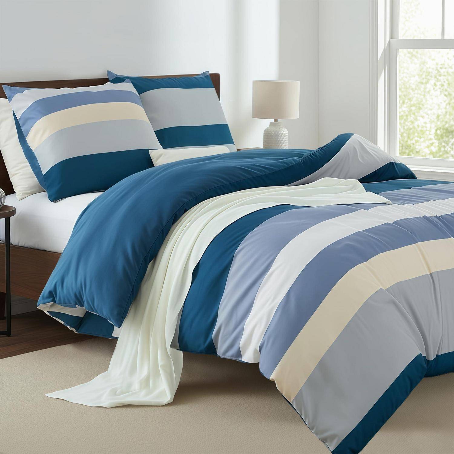 Blau-, weiß- und graugestreiftes Bettzug auf einem ordentlich gemachten Bett in einem hellen Schlafzimmer mit einer Nachttischlampe und Fenster.