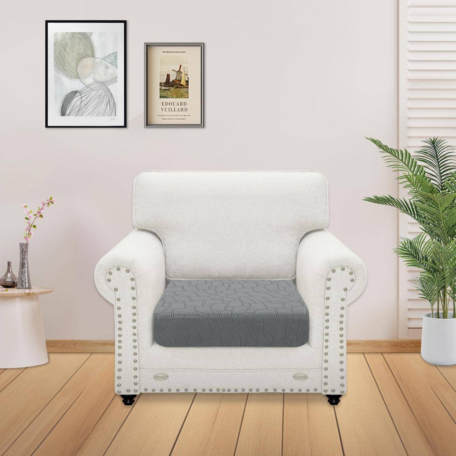 Hellbeiger bezogener Sessel mit grauem Sitzkissen in einem hellen minimalistischen Raum, flankiert von gerahmter Kunst und Topfpflanzen.