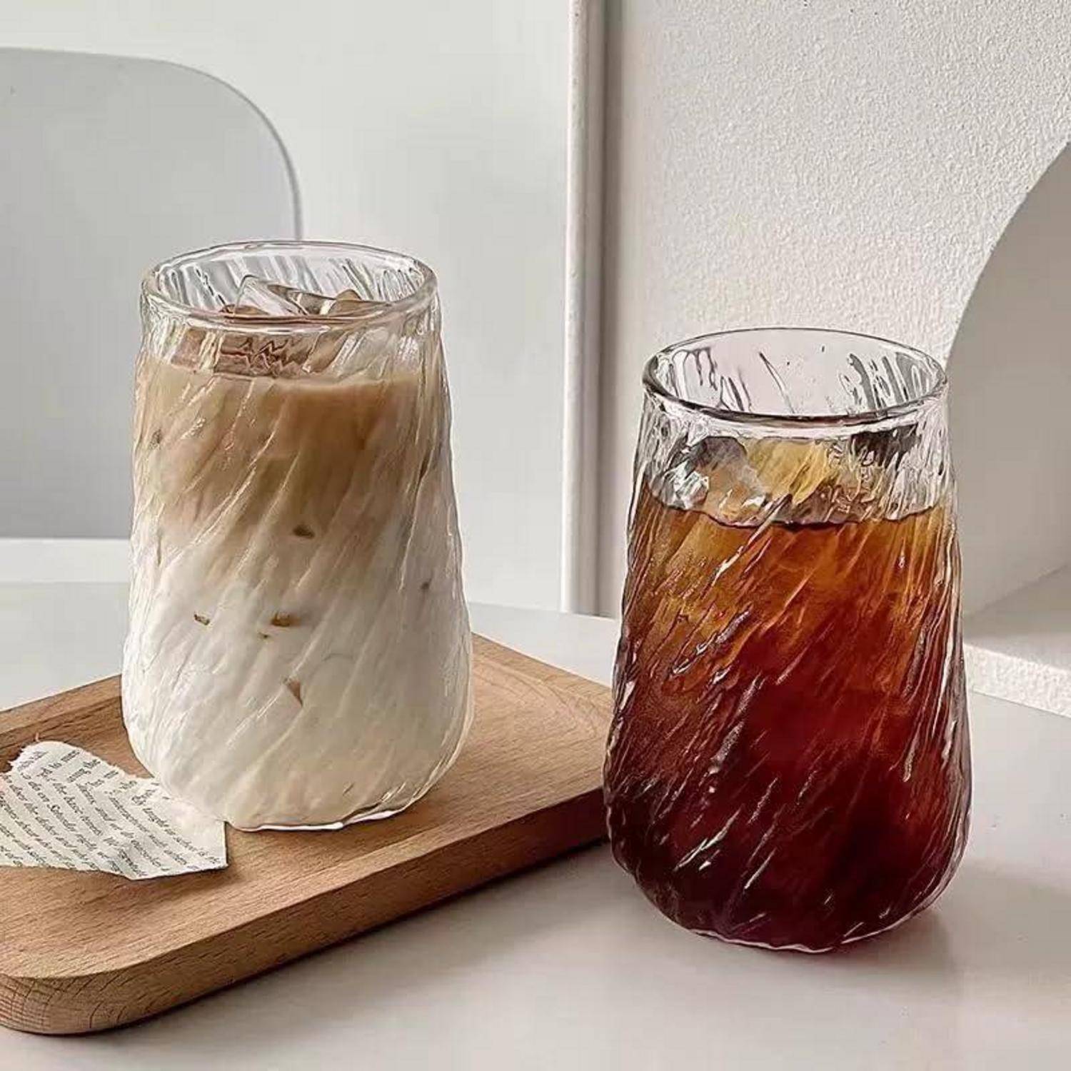 Zwei eisgekühlte Getränke in strukturierten, klaren Gläsern auf einem Holztablett, ein cremiger Latte und ein dunkler Tee, auf einem hellen Tisch.
