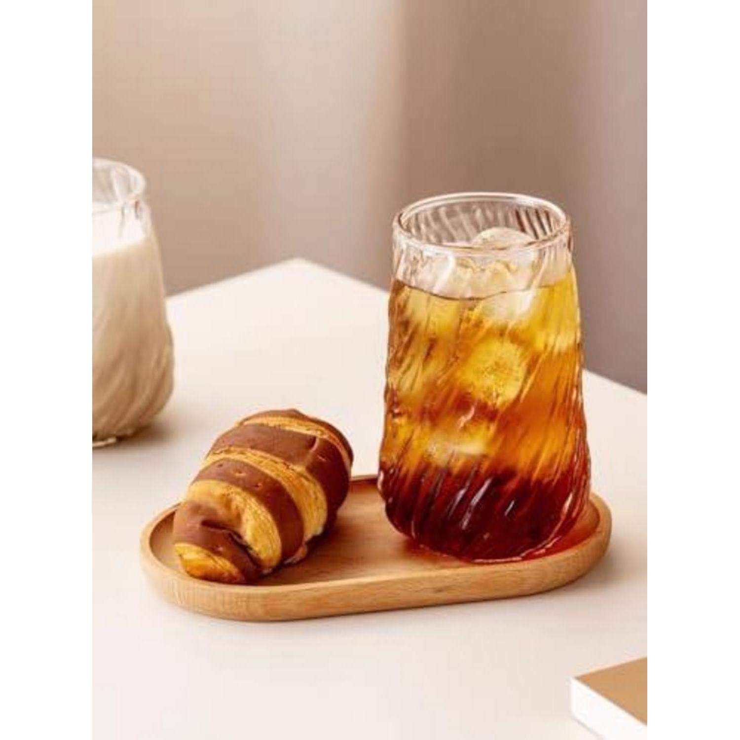 Glas mit eiskaltem bernsteinfarbenem Getränk und einem mit Schokolade beträufeltem Gebäck auf einem Holztablett auf einem weißen Tisch