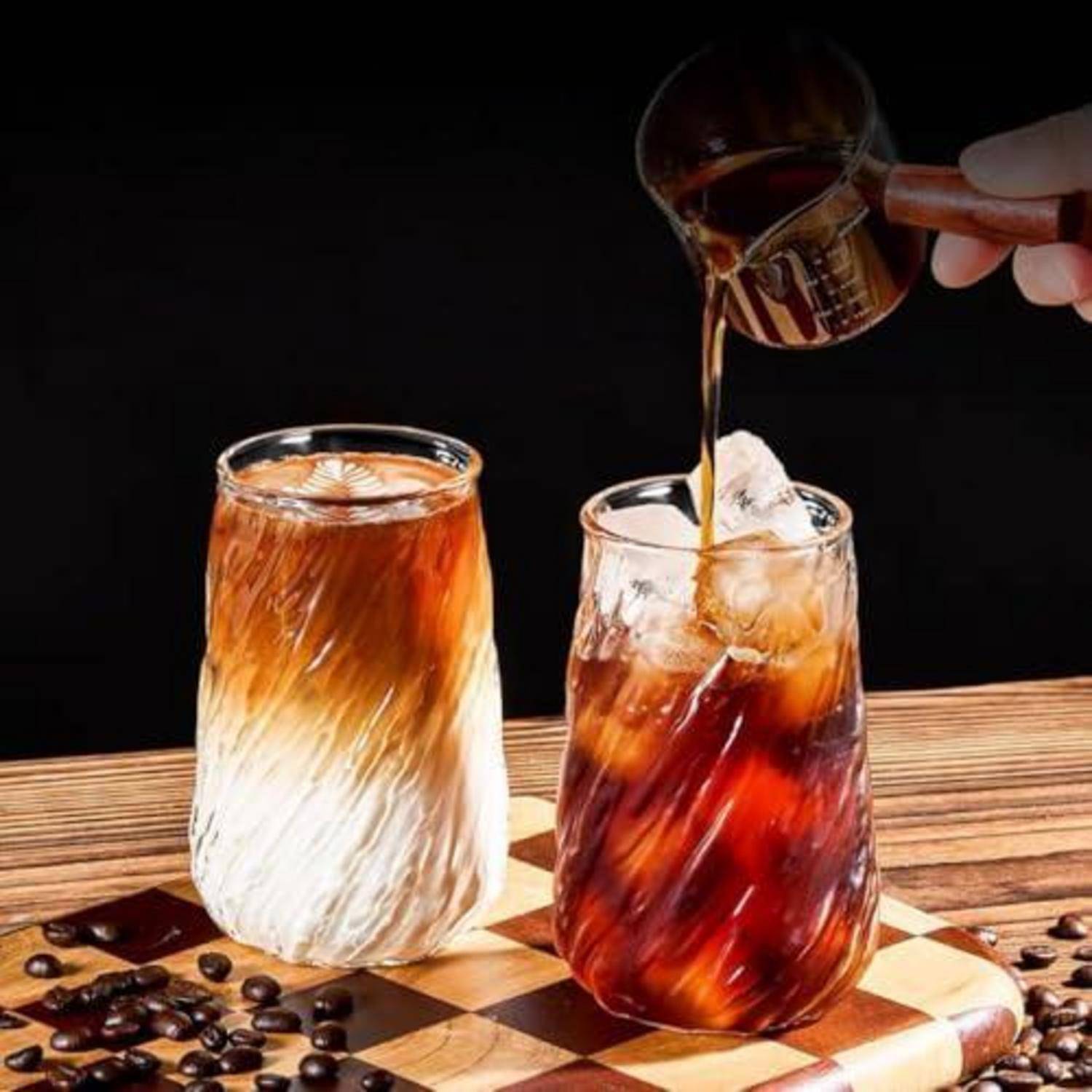 Eine Hand gießt Kaffeesirup in ein Glas Eiskaffee neben einem fertiggestellten Eiskaffee auf einem hölzernen Schachbrett mit verstreuten Kaffeebohnen.