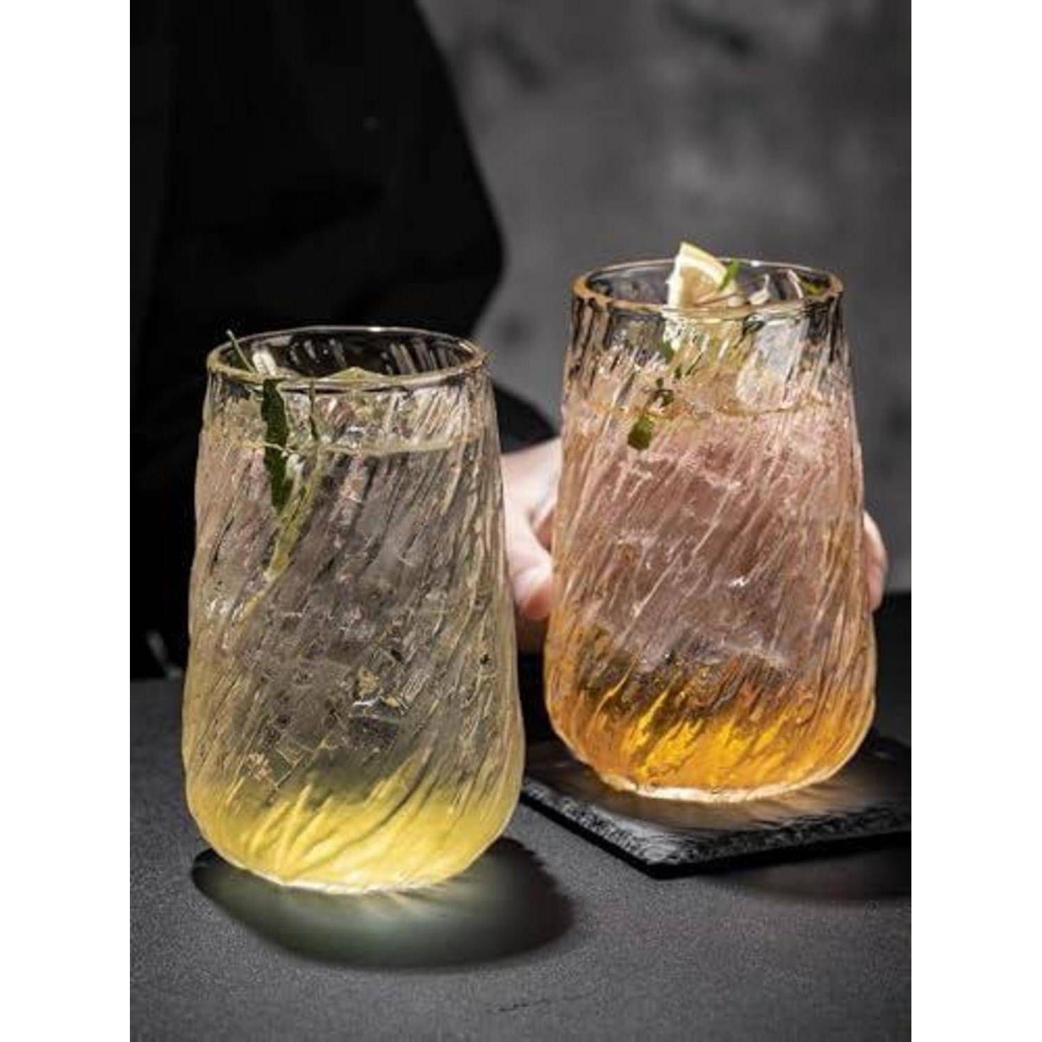 Zwei eisgekühlte Cocktails in strukturierten Gläsern auf einem dunklen Tisch, einer in Blassgelb und einer in Bernsteinfarbe, beide garniert mit Kräutern und Zitrusfrüchten.
