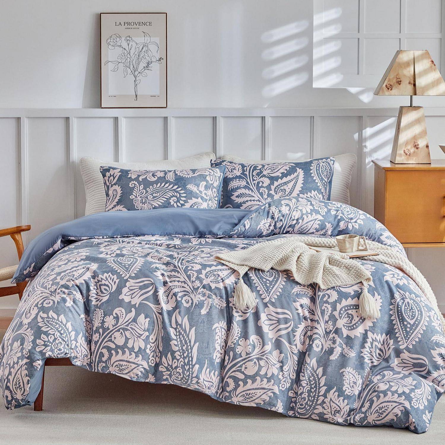 Ein ordentlich gemachtes Bett mit blauem Paisley-Bettwäsche und Kissen in einem hellen, modernen Schlafzimmer, mit einem gerahmten botanischen Druck und einer Nachttischlampe.