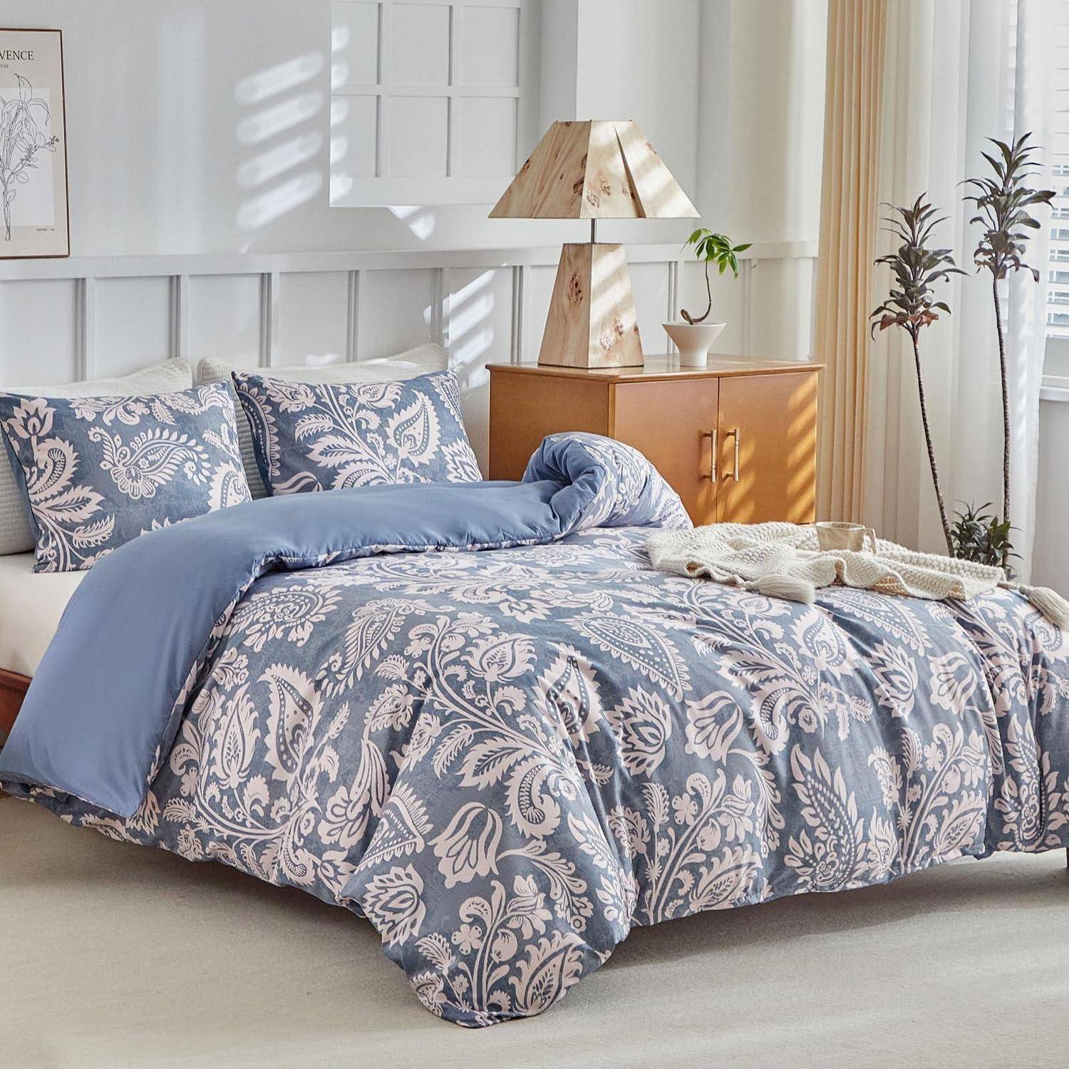 Ein ordentlich gemachtes Bett mit blaugrauem Musterbezug und passenden Kissen in einem hellen Schlafzimmer, neben einer hölzernen Kommode und einer Lampe.