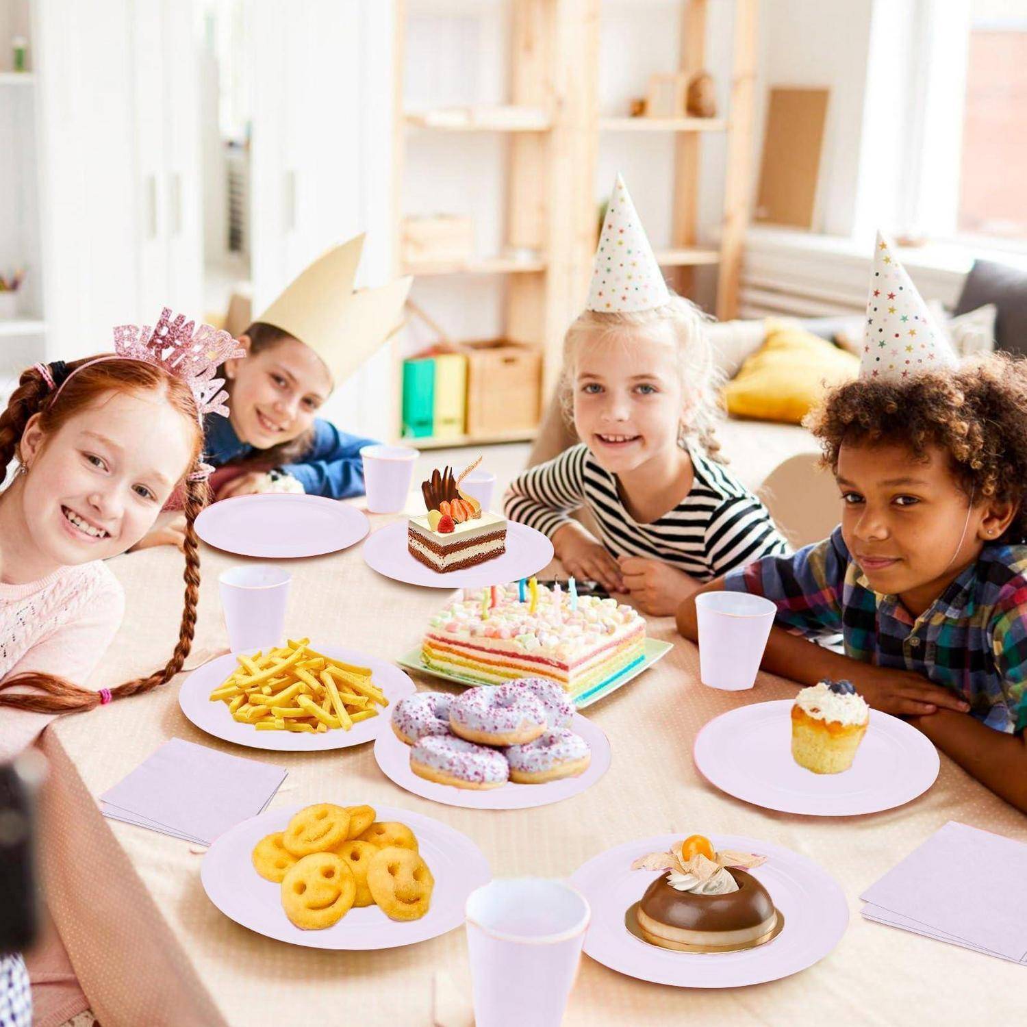 Kinder mit Partyhüten lächeln um einen Geburtstagstisch mit Kuchen, Cupcakes, Keksen, Pommes und Getränken in einem hellen Raum.