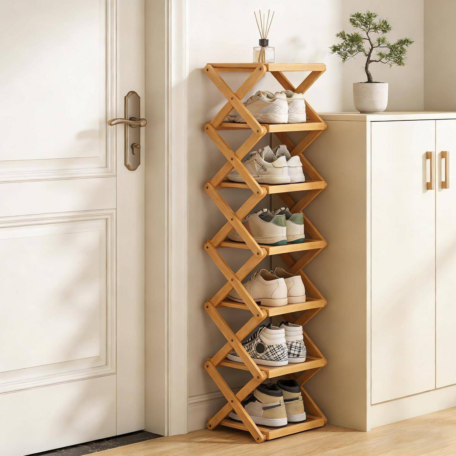 Hölzerner Schuhregal zum Zusammenklappen mit mehreren Schuhpaaren neben einer weißen Tür und einem Schrank mit einer Topfpflanze oben darauf.