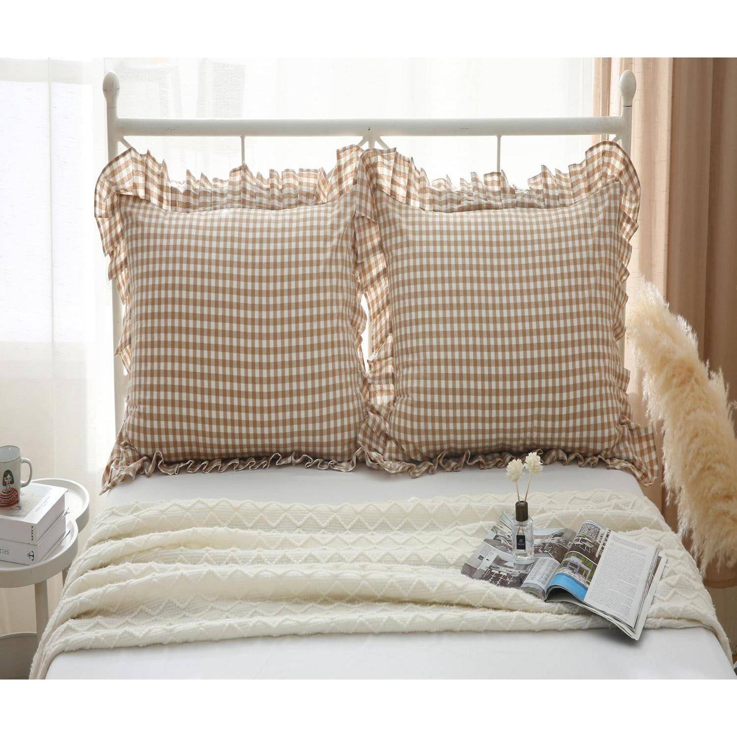 Ein ordentlich gemachtes Bett mit zwei braunen Gingham-Rüschenkissen, einer cremefarbenen Steppdecke und einer offenen Zeitschrift mit einer kleinen Vase obenauf in einem hellen Zimmer.