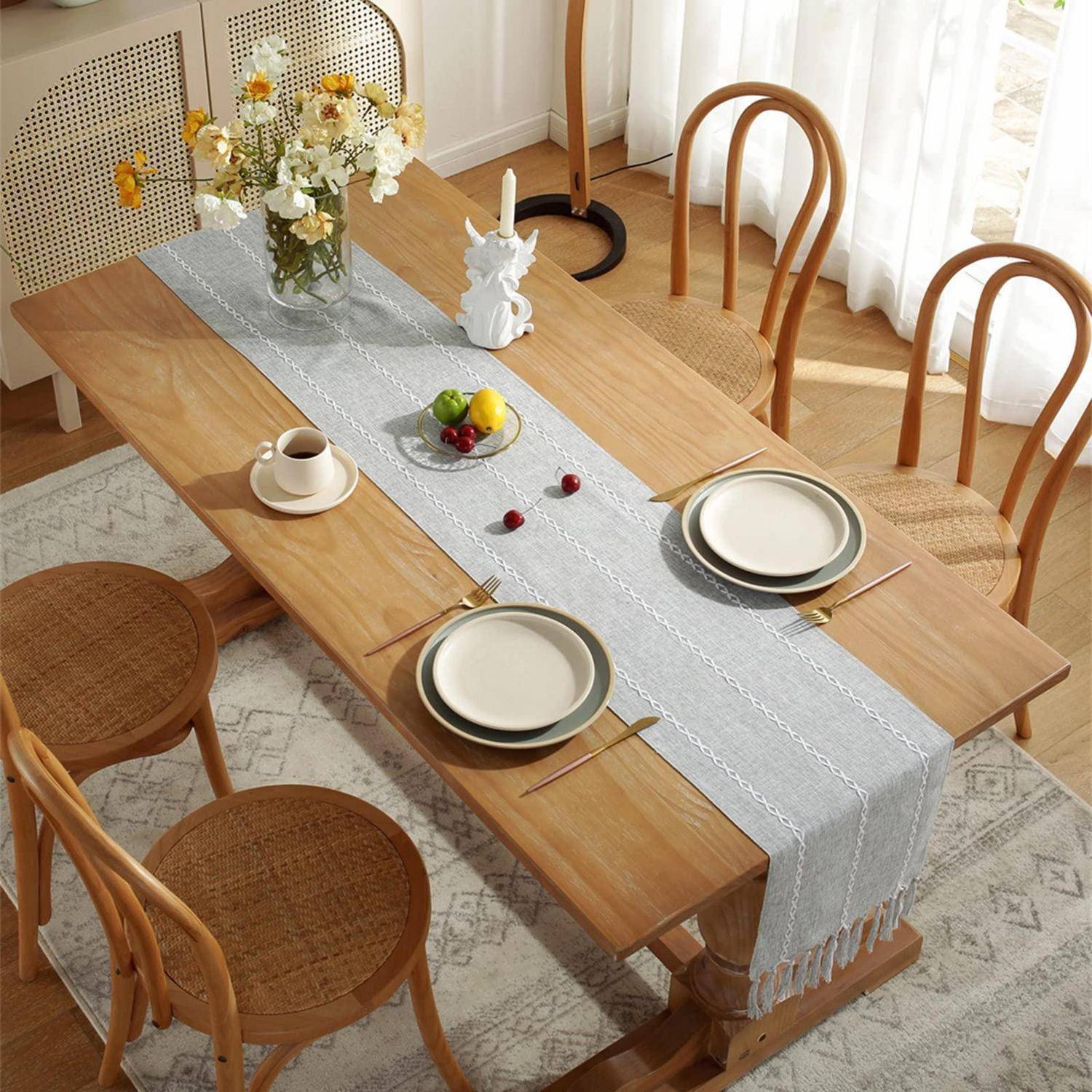 Ein Holzesstisch für vier Personen mit Tellern, Tassen, Obst, Blumen und einem grauen Tischläufer in einem hellen Raum.