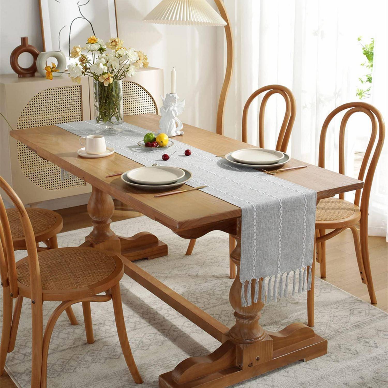 Esstisch für zwei Personen in einem hellen Raum eingedeckt, mit Holztisch, beigen Stühlen, Tellern, Tassen und einer Blumenvase auf einem Tischläufer.