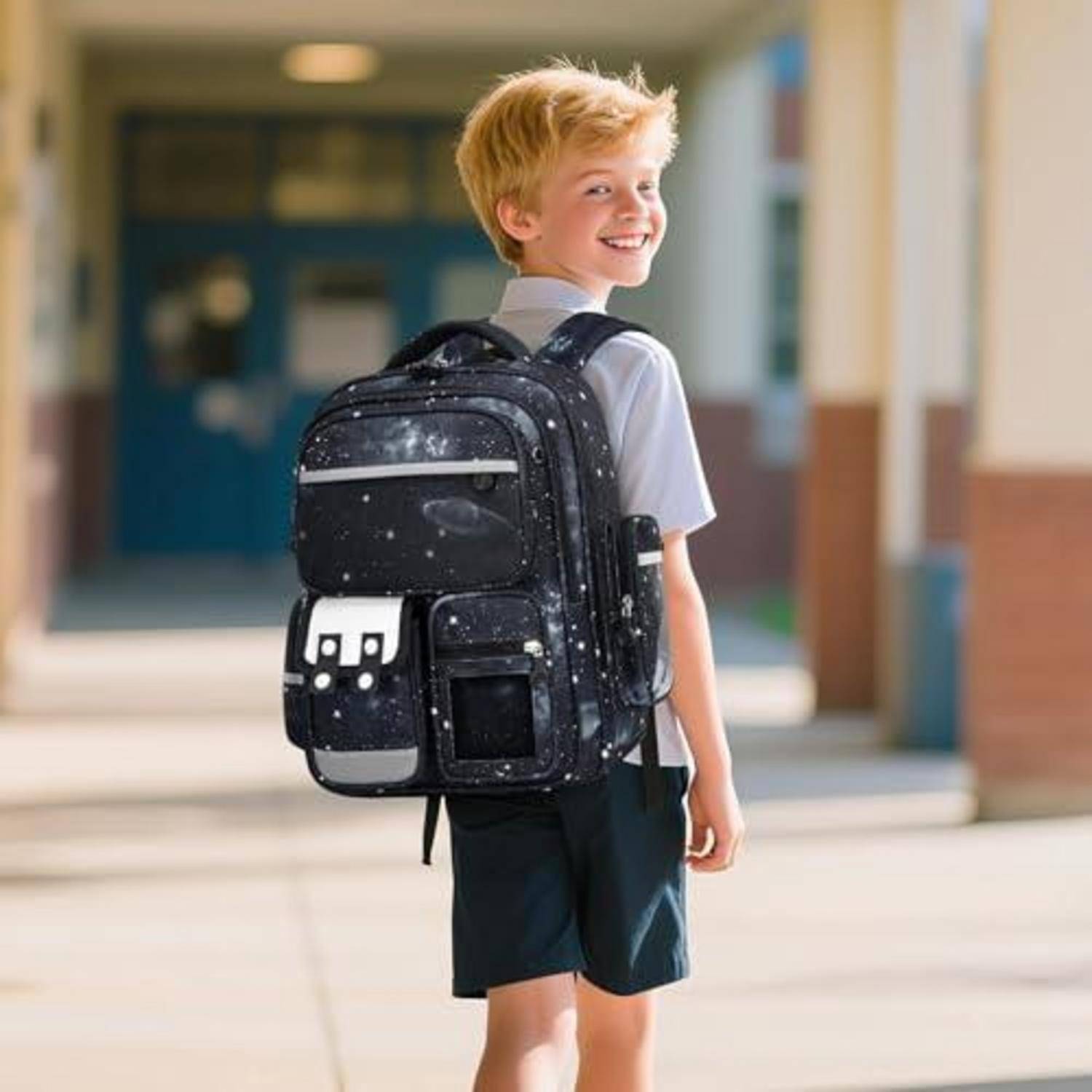 Lächelnder junger Junge mit einem großen schwarzen Rucksack, der außerhalb eines Schulflurs steht und bereit für den Unterricht ist.