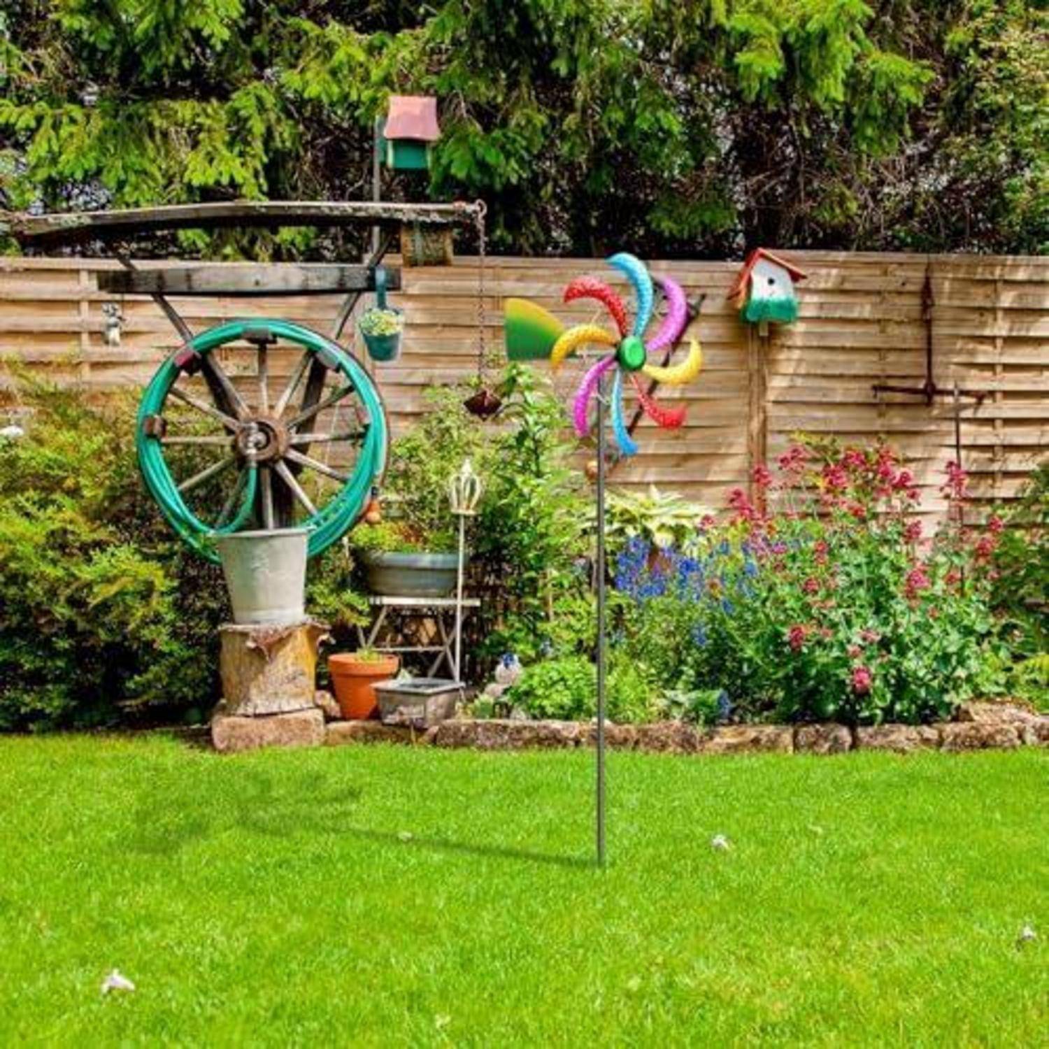 Farbenfroher Garten mit einem regenbogenfarbigen Windrad, einem alten Wagenwrad-Pflanzkübel, Blumenbeeten und einem Holzzaun im Hintergrund.