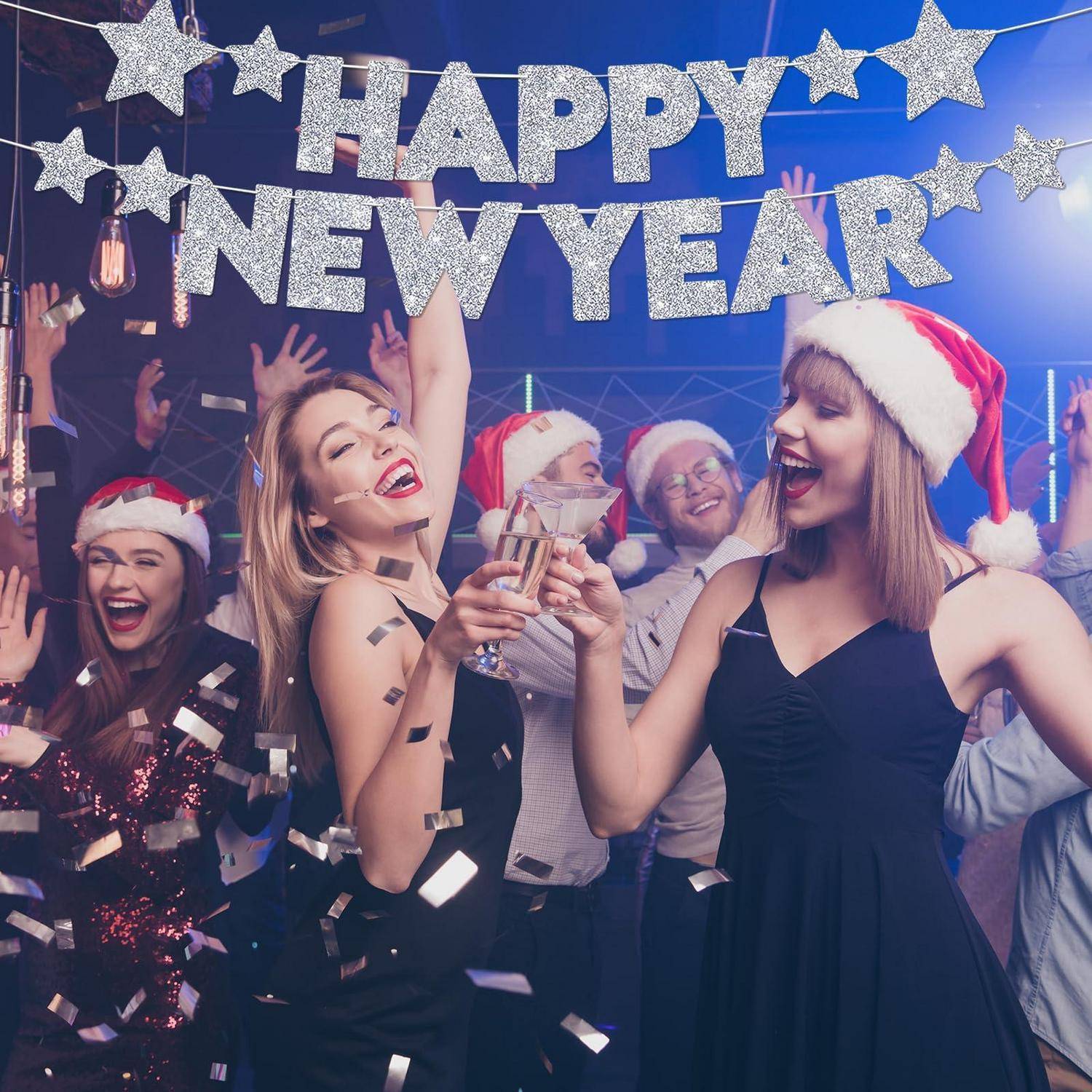 Feiernde in Weihnachtsmützen stoßen mit erhobenen Gläsern unter einem „HAPPY NEW YEAR"-Banner und fallenden Konfetti auf das neue Jahr an.