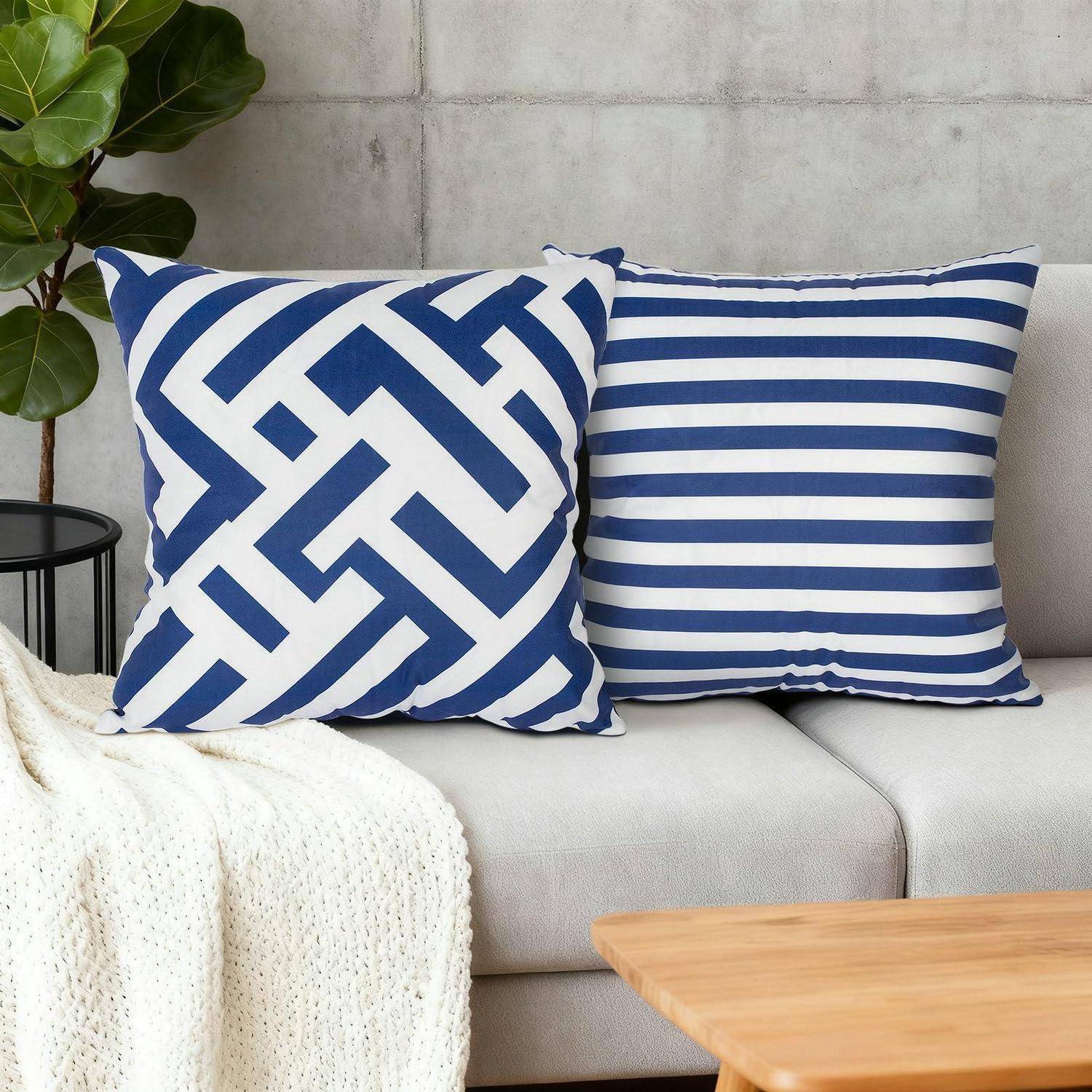 Zwei blau-weiß gemusterte Wurfdekokissen auf einem grauen Sofa, eines mit geometrischem Muster und eines mit Streifen, neben einer Pflanze und einer Decke in einem Wohnzimmer.
