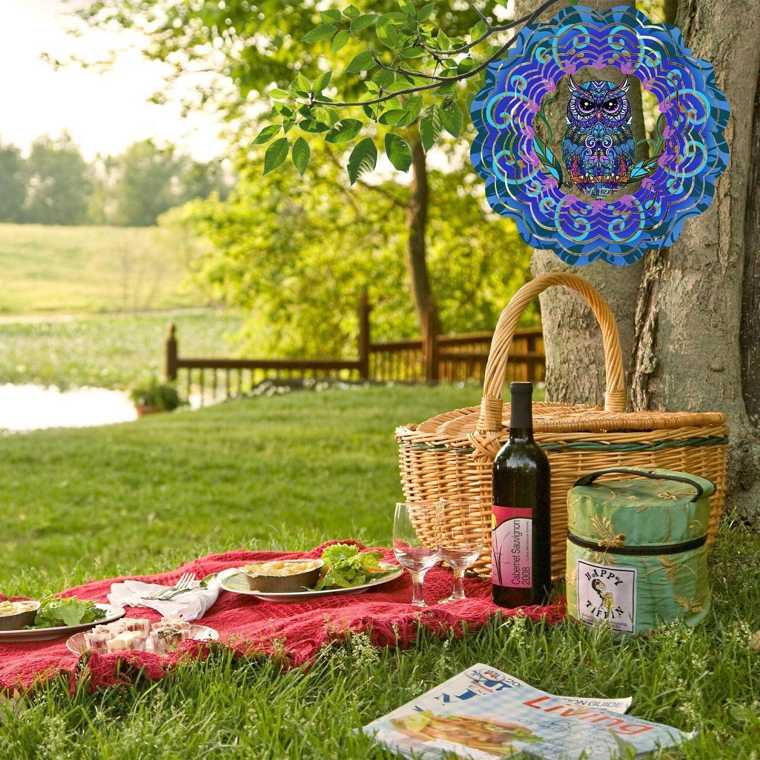 Picknick-Setup auf dem Gras unter einem Baum mit Korb, Weinflasche, Gläsern, Tellern mit Speisen und einer Zeitschrift; dekorative Eule Windspiel darüber.