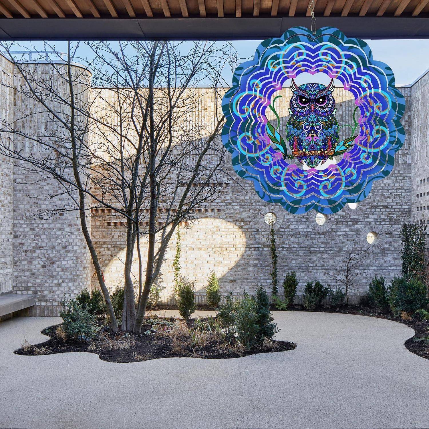 Außenhof mit einem Baum und bepflanztem Blumenkübel, mit einer großen, bunten Eule im Bleiglasfenster-Stil als hängende Skulptur vor einer Steinmauer.