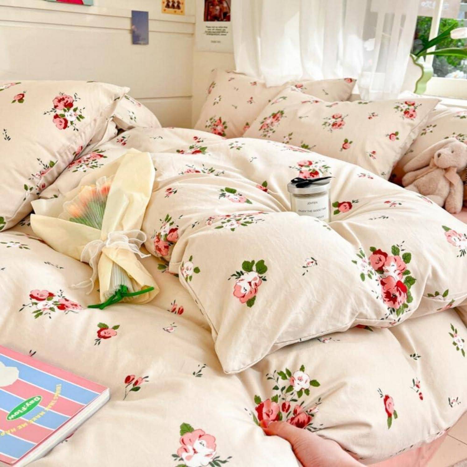 Gemütliches Bett mit cremefarbener Blumenbettwäsche, einem Blumenstrauß, einer Kerze und einem Teddybär am Fenster in einem hellen Schlafzimmer.