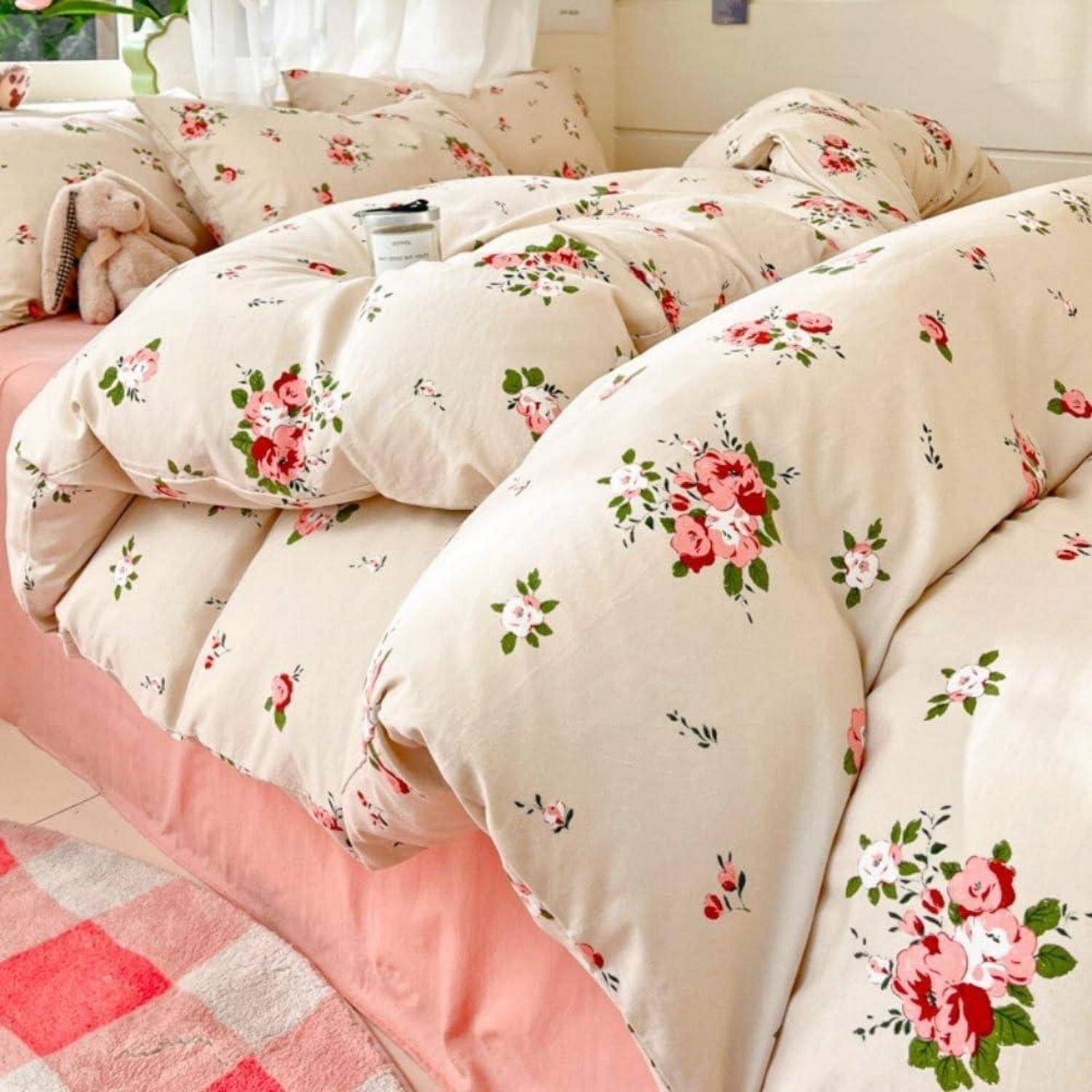 Bett mit einer cremefarbenen Blumensteppdecke und Kissen, mit einem rosa Bettrock und einem rosa karierten Teppich in einem hellen Schlafzimmer.